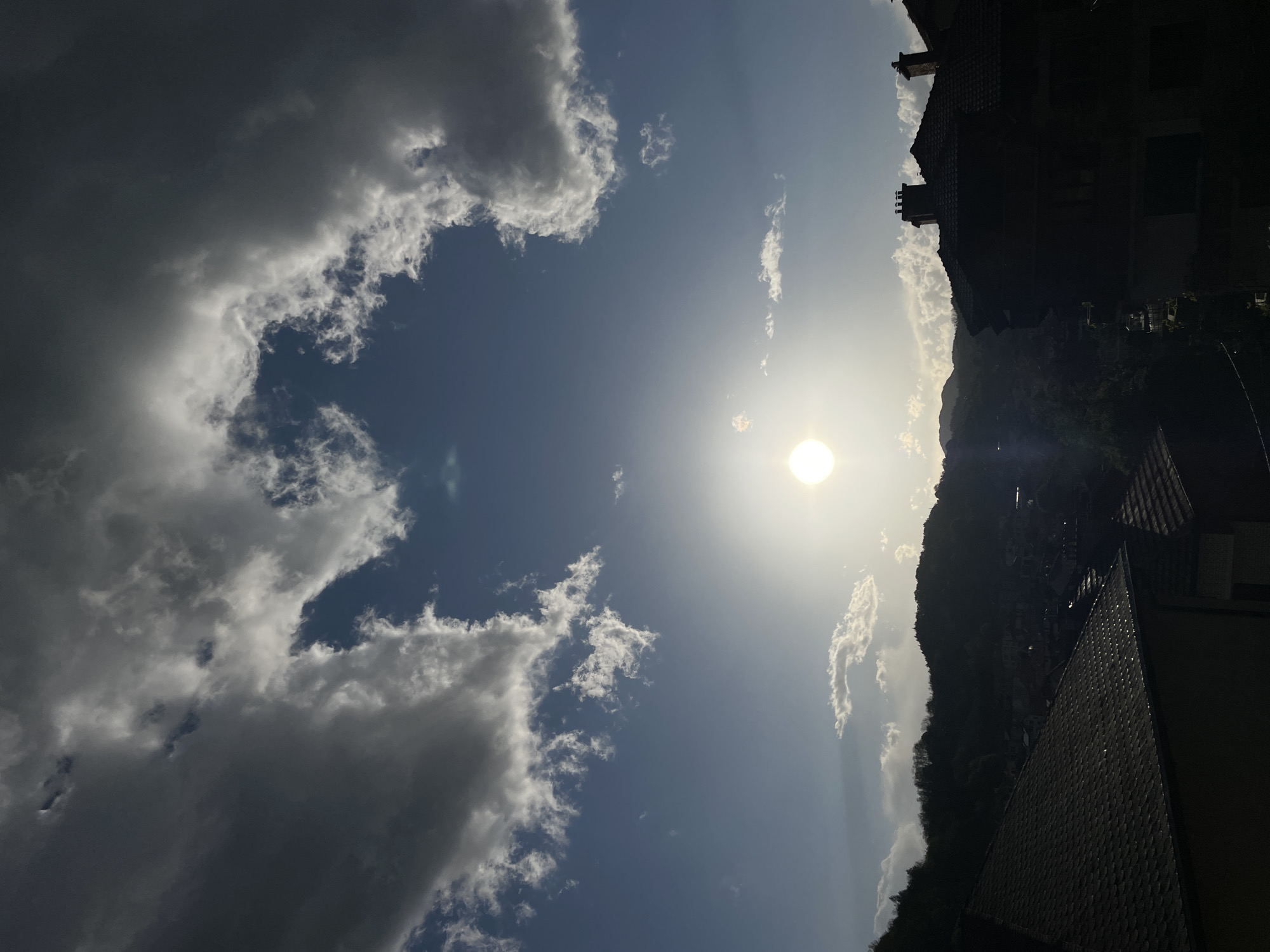 The image shows a view in Zlatograd, Bulgaria, with a bright sun obscured by haze on the right side. To the left is a blue sky with fluffy white and gray clouds. On the right, there are silhouetted buildings, trees, and a hillside. A building is visible in the foreground with a tiled roof.