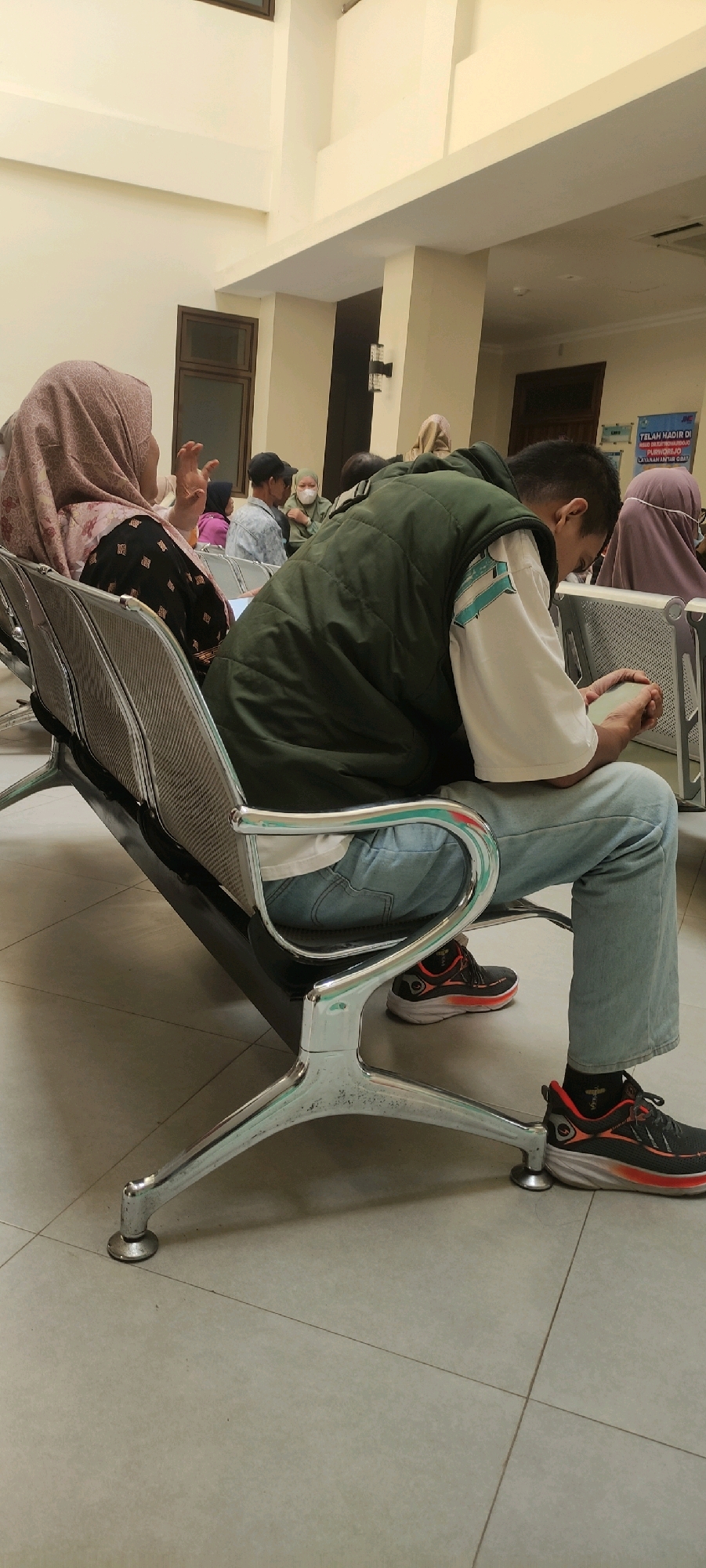 In Purworejo, Indonesia, at what appears to be a waiting area, possibly a clinic or hospital, several individuals are seated on metallic chairs with mesh backs and chrome frames. The floor is tiled in a light color, and the walls are painted a pale yellow.
In the foreground, a young person wearing a dark green vest over a white t-shirt with a teal graphic is engrossed in a mobile phone. They are dressed in light blue jeans and dark athletic shoes with red accents and white soles.
To the left, a woman wearing a patterned pink hijab and a black patterned dress is gesturing with her right hand while facing away from the camera. She is seated on a similar metallic chair. Other individuals are visible in the background, some wearing masks, suggesting a public health setting. One person in the background is wearing a light green hijab and a matching garment, and another is wearing a light blue patterned shirt. A poster with text, partially obscured, is visible on the wall in the far background.
