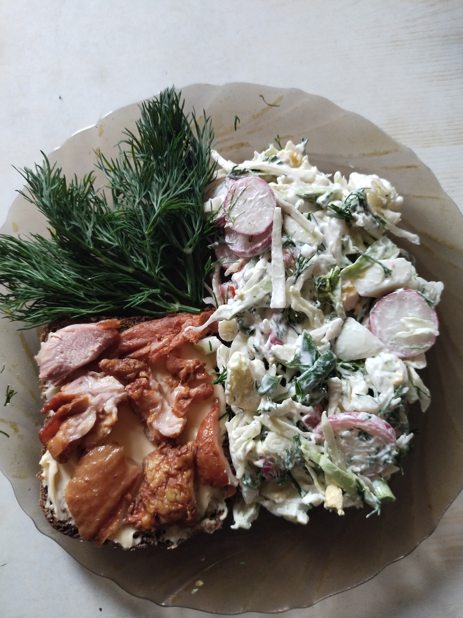 An overhead shot captured in Horishni Plavni, Ukraine, displays a single serving of food arranged on a translucent, light brown, wavy-edged glass or plastic plate. The plate rests on a light-colored surface, possibly wood or laminate, exhibiting a subtle grain pattern. On the left side of the plate, a slice of dark bread is visible, topped with a light-colored creamy spread and an assortment of sliced meat. The meat includes lighter, pinkish pieces, possibly cooked poultry breast, alongside darker, irregular, and more textured pieces that appear to be smoked or fried chicken skin or dark meat. A substantial bundle of fresh green dill sprigs is placed across the top of the meat, extending onto the salad. The right side of the plate holds a creamy salad composed of shredded white cabbage, thinly sliced pink and white radishes, chopped green herbs (likely more dill and/or green onion), and discernible pieces of hard-boiled egg. The salad is thoroughly coated in a thick, white, creamy dressing, indicative of a mayonnaise-based preparation. No individuals are present in the scene.