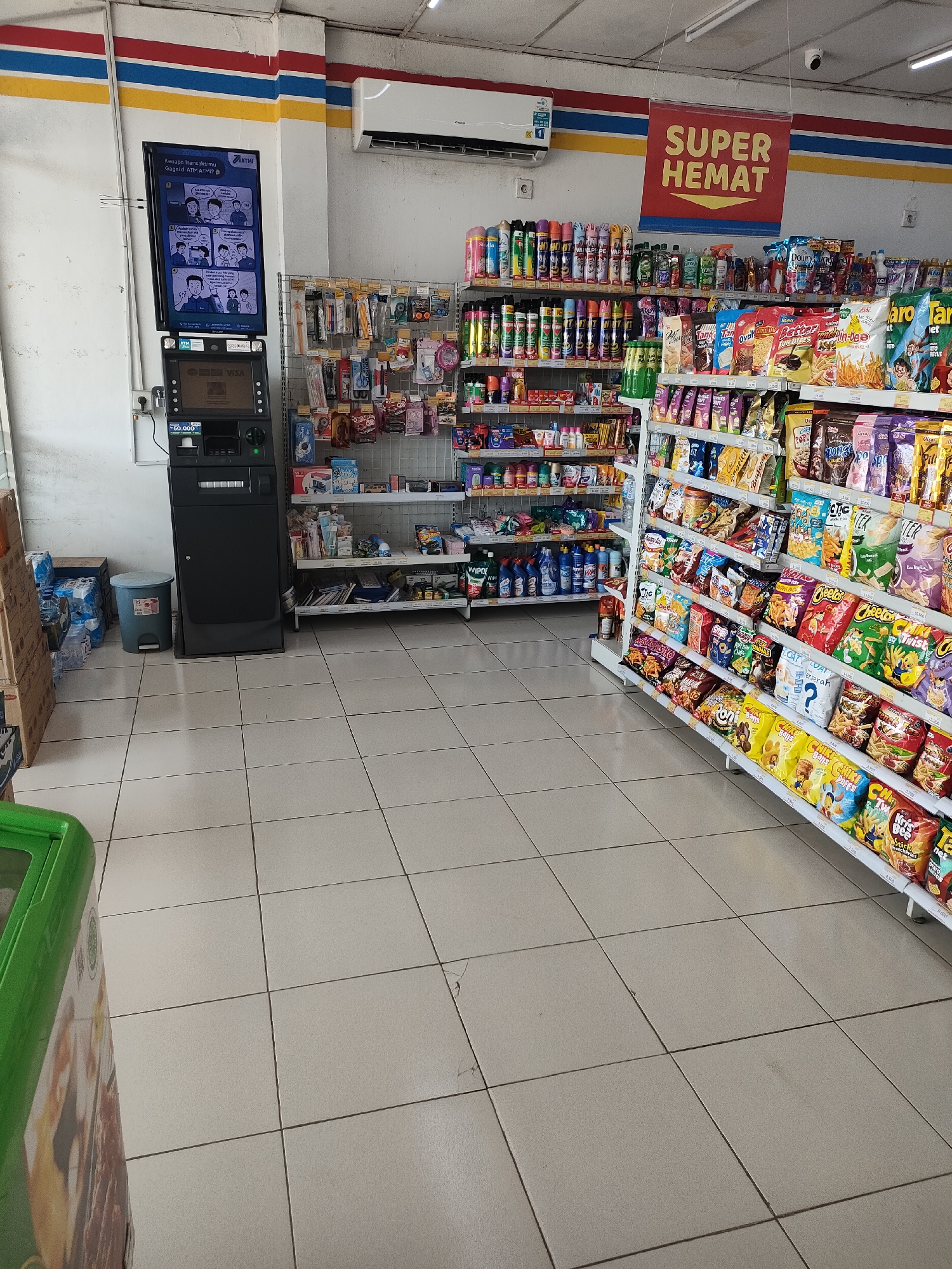 The image depicts the interior of a convenience store, possibly a minimarket, in Curahdringu, Indonesia. The store is well-lit and stocked with various products. In the foreground, on the left, a green-colored freezer unit is partially visible. The floor is tiled in a light beige color.
Dominating the mid-ground and background are rows of shelves filled with merchandise. On the left side, there is an ATM machine with a digital screen displaying information. Adjacent to the ATM, there are stacks of bottled water and a blue trash can. Further to the left, on the wall, there are several rows of shelves containing cleaning supplies and personal care items.
The right side of the store features numerous shelves packed with packaged snacks and drinks. These shelves are organized and display a wide variety of colorful products. A prominent red banner with yellow text reading "SUPER HEMAT" (meaning "Super Economical" or "Great Savings") is hung from the ceiling, indicating a sale or promotion. Above the shelves, an air conditioning unit is visible on the wall. The overall impression is that of a typical, busy retail environment. There are no people visible in the image.