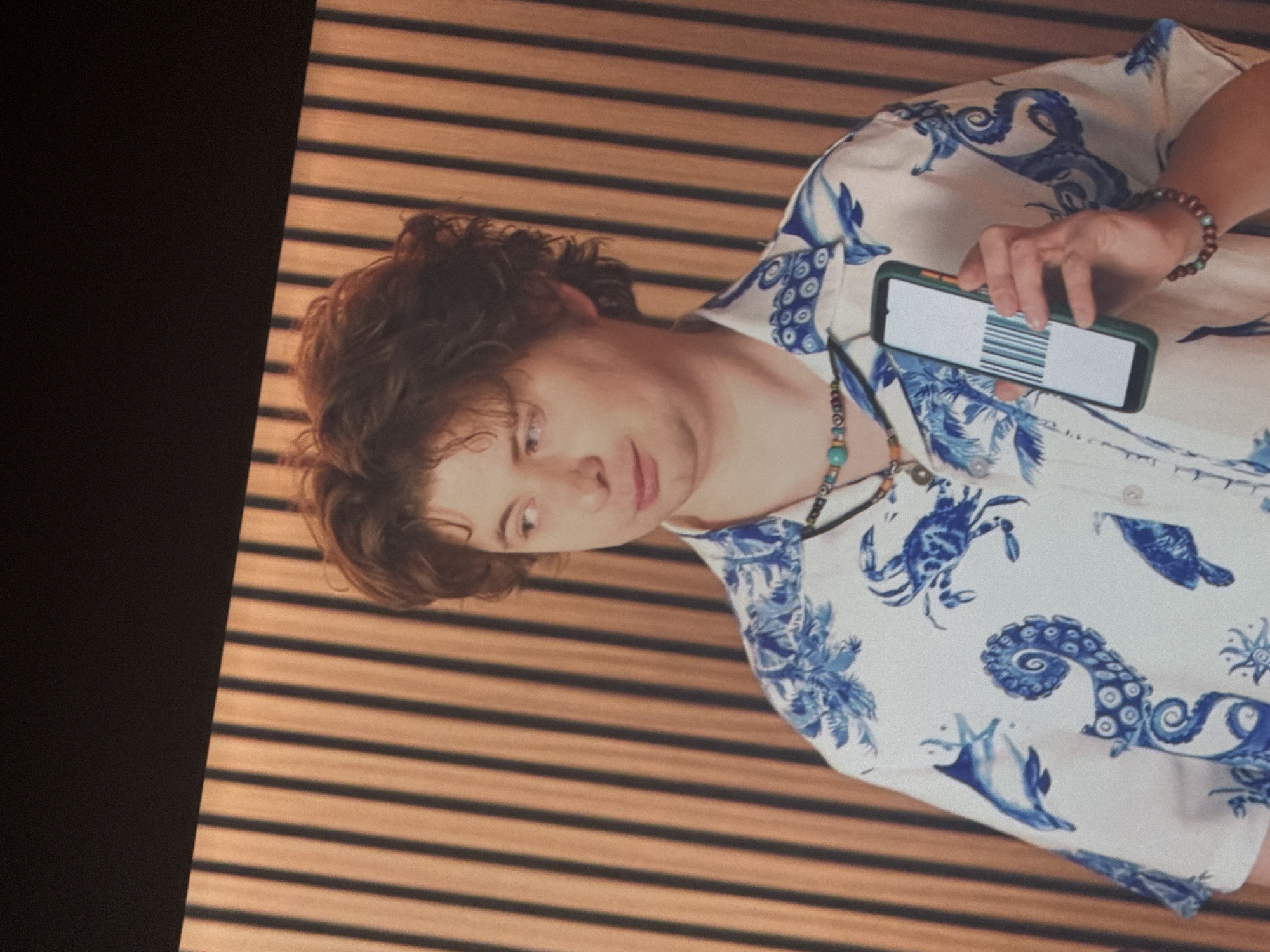A young man with reddish-brown curly hair is standing in front of a wood-paneled wall. He is wearing a white short-sleeved button-up shirt with a blue pattern of marine life, including crabs, fish, and octopuses. He is also wearing a beaded necklace and a beaded bracelet. In his right hand, he is holding a smartphone with a barcode displayed on the screen. The man is looking off to his left. The overall lighting suggests an indoor setting.