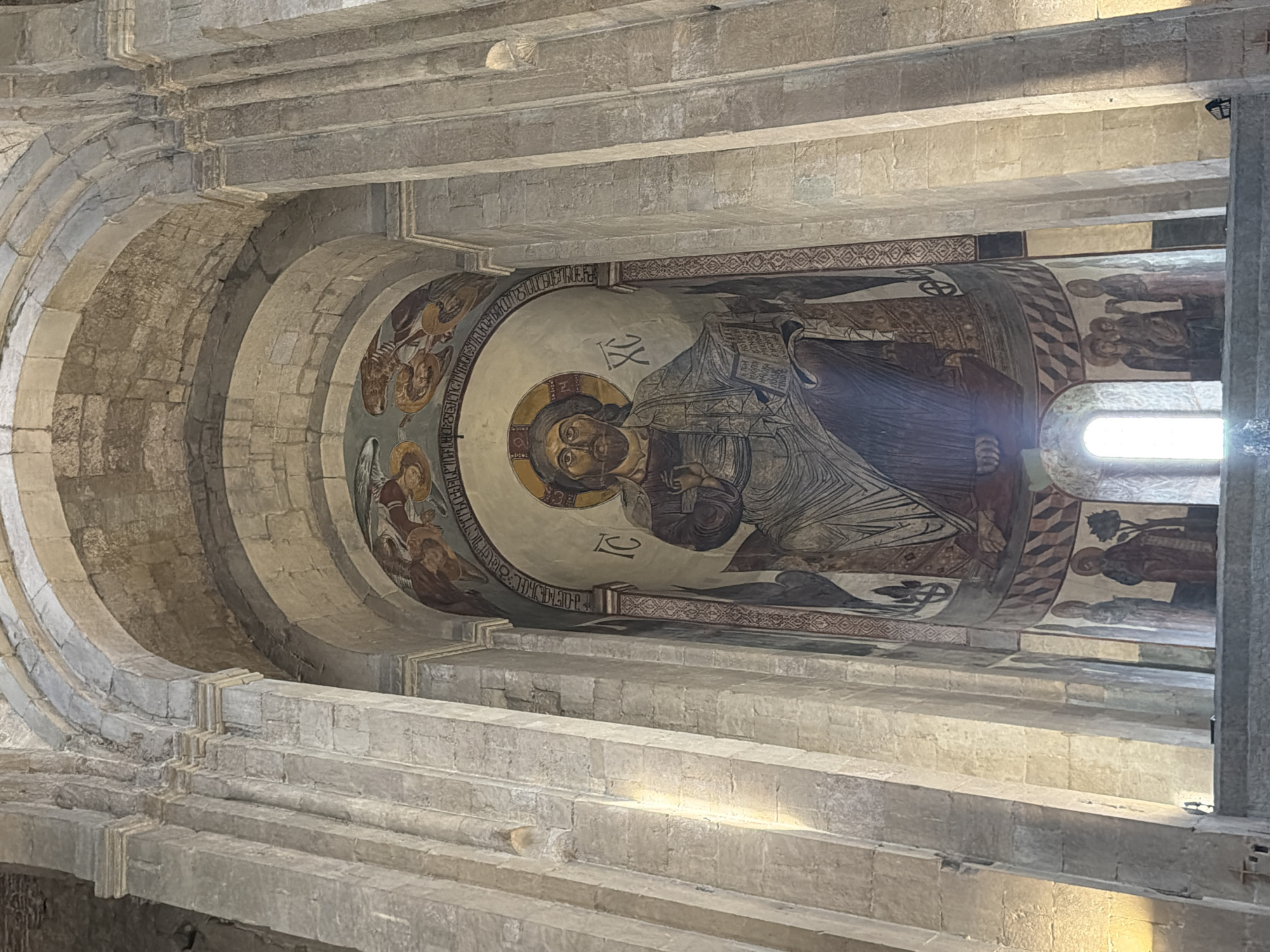The image captures a section of the Svetitskhoveli Cathedral in Mtskheta, Georgia, specifically focusing on the mural art located within the structure.
The camera is looking up at a painted mural which is located inside an alcove. The mural depicts a central figure with a halo, robes, and a book. Other figures, some possibly angels, are arranged around the central one. Decorative borders and writing are around the primary image.
The architecture features stone arches, layers of stone bricks, and window. The lighting is a mix of natural sunlight coming through the window and general ambient light.