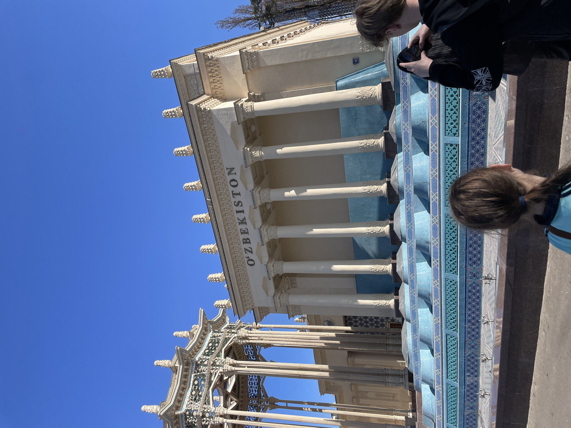 The image depicts a low-angle view of the Uzbekistan pavilion at the All-Russia Exhibition Center (VDNKh) in Moscow, Russia. The building features white columns with ornate detailing and blue accents. The name "O'ZBEKISTON" is written in white, capitalized letters above the columns. The sky is clear blue.
Two people are partially visible in the foreground, on the right. One person is looking at a mobile phone, and another person is facing away from the camera. A multi-tiered structure with detailed lattice work is visible on the lower left, appearing to be a gazebo or similar architectural feature related to the pavilion. There are outdoor water fountains or water features in the foreground.