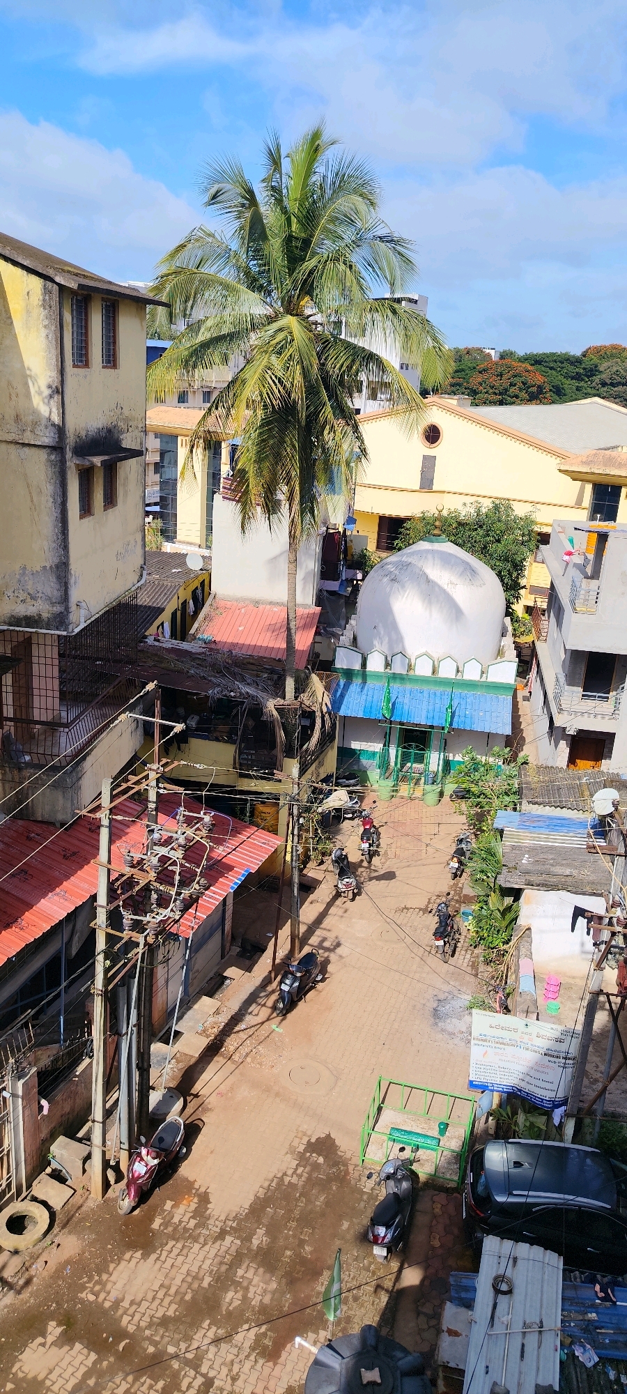 This image captures a street scene in Belagavi, India, characterized by a mix of residential buildings, a mosque, and utility infrastructure. A prominent coconut palm tree stands tall in the center, its fronds reaching towards a blue sky dotted with white clouds. The street below is paved with interlocking bricks and shows signs of recent moisture, creating reflective puddles.
Several parked motorcycles and scooters are visible on the street, indicating a common mode of transportation in the area. To the right of the street, a mosque with a white dome and green accents is a central structure. Adjacent to the mosque, a small, enclosed green structure houses what appears to be a grave marker.
On the left side of the image, a multi-story building with a yellow facade and windows is present. Electrical poles with numerous wires and transformers are strung across the scene, highlighting the urban utility network. In the background, more buildings and greenery can be seen, suggesting a densely populated urban environment. A black car is parked near the mosque. A banner with text in a South Indian language is affixed to a wall on the right side of the frame. The overall impression is of a bustling, lived-in urban environment under a bright, sunny sky.