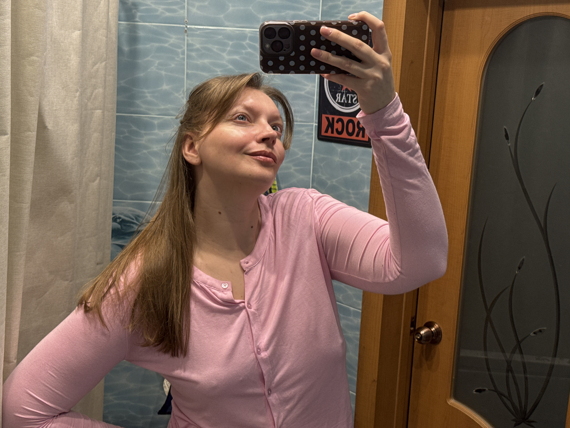 A fair-skinned woman with light brown hair is taking a selfie in a bathroom. She is wearing a long-sleeved pink cardigan. The background includes blue tiled walls, a shower curtain or partition, and a wooden door with a glass insert featuring a dark floral design. There is also a "Rock Star" sign visible on the wall behind her. The image doesn't provide sufficient information to confirm its location in Izhevsk, Russia.