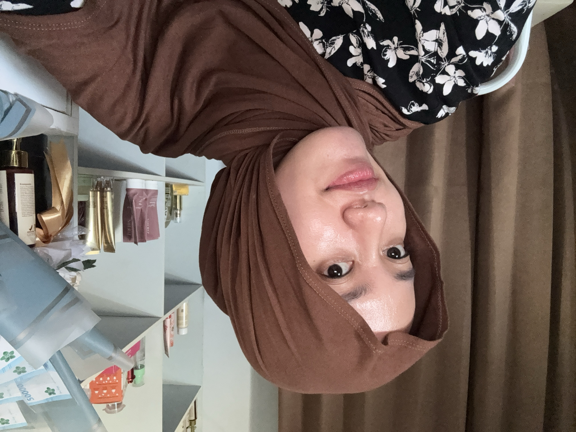 A woman wearing a brown headscarf and floral print top is indoors. Shelves with various cosmetic and personal care products are to the left of the frame, while a brown curtain is on the right. The location is potentially Gadingrejo, Indonesia, but there is no specific evidence in the image to confirm this.