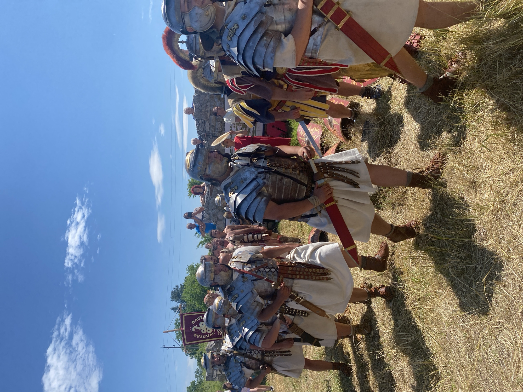 The image depicts an outdoor historical reenactment in Gigen, Bulgaria, under a clear blue sky with scattered white clouds. The foreground and mid-ground are dominated by approximately eight individuals dressed as Roman legionaries, wearing lorica segmentata (segmented plate armor), helmets (galea), white tunics, and brown caligae (sandals or boots). Each carries a gladius (short sword) in a scabbard, and several have a cingulum militare (belt with hanging straps). They are walking in a loose formation across a field of dry, light-brown grass, casting distinct shadows indicating bright sunlight.
In the mid-ground to the left, a maroon banner with yellow text, partially visible, displays "LEG IIII FLAVIAE NAVT". This banner is affixed to a wooden pole. Behind the legionaries, several red, oval scuta (Roman shields) with gold decorative elements are propped on the ground.
In the mid-ground, behind the reenactors, a group of spectators observes from atop a stone wall or ruin. Some are actively watching, while others appear to be photographing with mobile phones. Further back, more costumed individuals are visible, including one person in a red garment holding a fan on the left, and a figure with a red-plumed helmet on the far right. The background features green trees, a utility pole with power lines, and distant structures, under the bright sky.