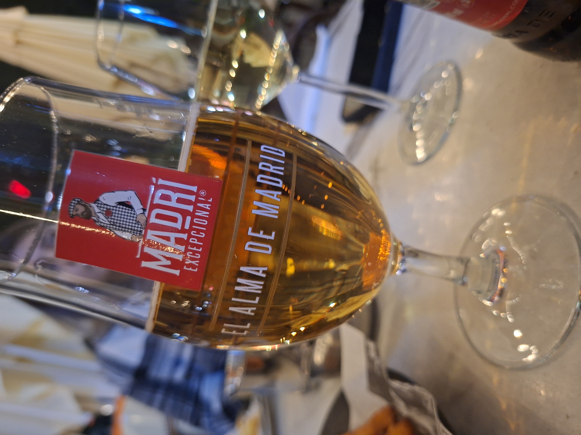 The image shows two glasses, one filled with a golden liquid, likely beer, and another with a lighter-colored liquid, possibly white wine. The beer glass has a red label with "Madrid Excepcional" written on it. The scene is indoors, possibly in a restaurant or bar. The location is implied by the presence of the beer glass, with the label “El Alma de Madrid,” suggesting a connection to the city of Madrid. There is no information to determine the location to be Plovdiv, Bulgaria.