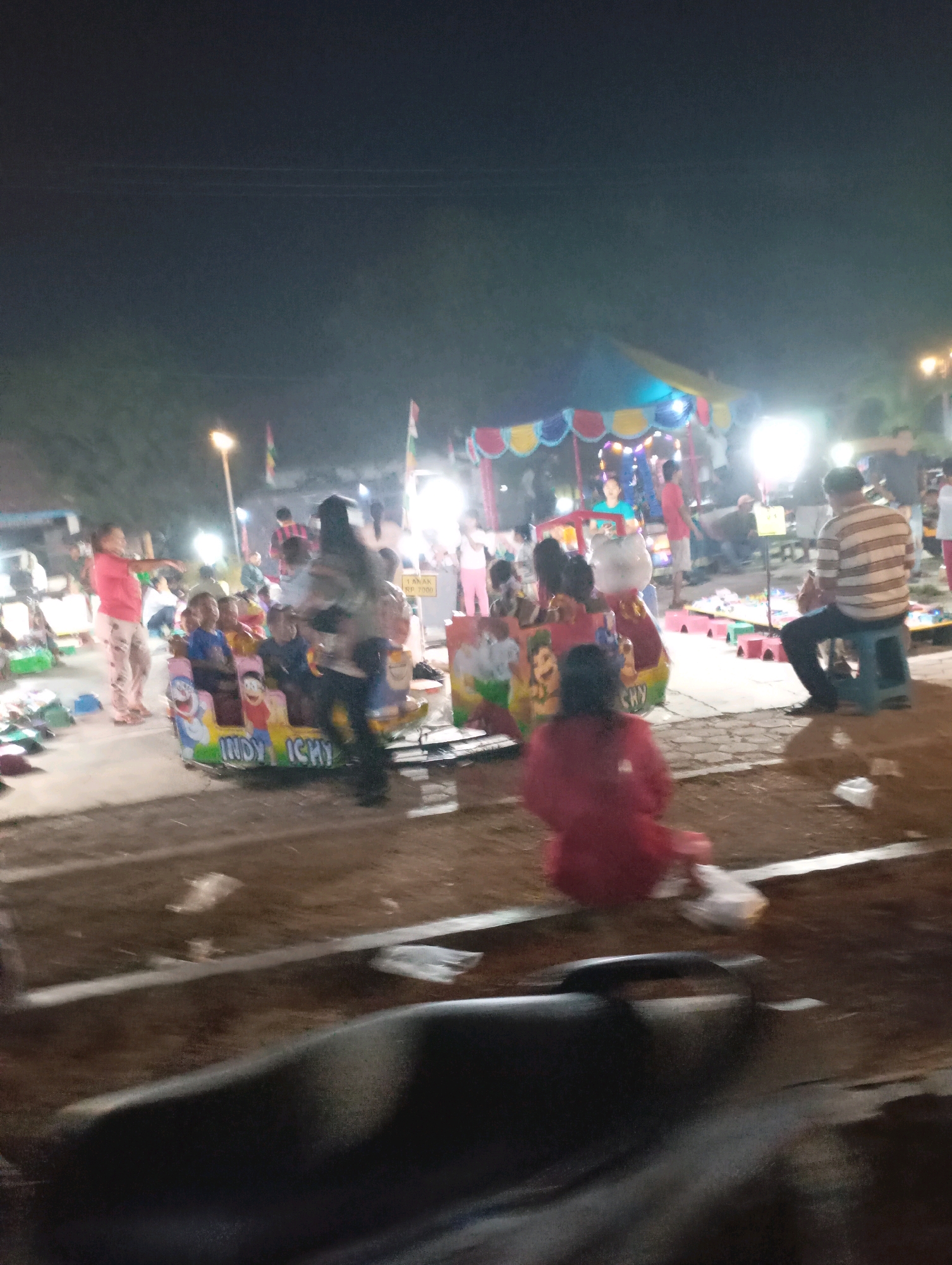 The image depicts a night scene at a local fairground, likely in Kecamatan Haurgeulis, Indonesia, as indicated by the context of the query. In the foreground, a blurry dark object, possibly a part of a vehicle or furniture, is visible. The middle ground is dominated by a small amusement ride with cartoon characters and the text "INDY ICHY". Several children and adults are present around the ride, with some children seated on it. A woman in a pink top and patterned pants stands to the left, gesturing. In the background, a brightly colored tent with lights is set up, suggesting a performance or game area. Various other people, stalls, and streetlights are scattered throughout the scene, illuminated by artificial lighting. The ground appears to be a mix of dirt and paved areas, with some litter visible. The overall atmosphere is lively and communal, typical of a local evening fair.