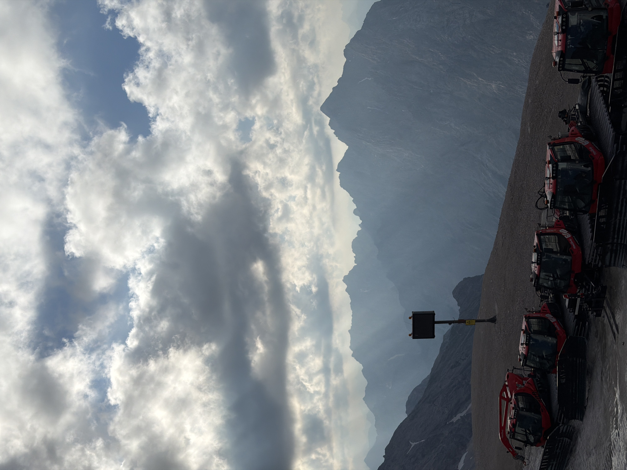 The image depicts a scenic view of the Bavarian Alps, likely from the Zugspitze, the highest peak in Germany, near Garmisch-Partenkirchen. In the foreground, several red snow groomers are parked on a dusty, gravelly surface. These machines are designed for preparing ski slopes. Behind the snow groomers, a dark, square signpost with two orange lights on top stands erect, accompanied by a yellow triangular warning sign below. The background is dominated by a vast mountain range, with peaks fading into a hazy blue atmosphere. Sunlight breaks through a dramatic, cloudy sky, casting rays that illuminate parts of the distant mountains. The overall scene suggests a high-altitude location, possibly a ski resort area during a break in operations or an off-season. There are no people visible in the image.