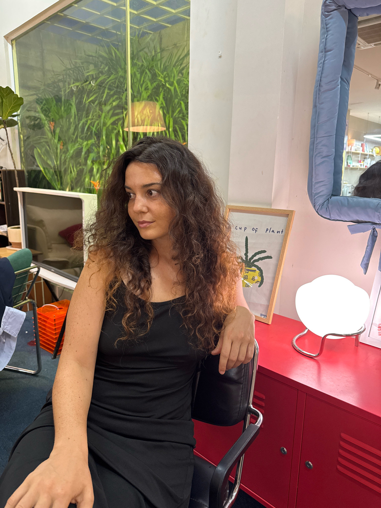 A woman with curly brown hair is seated in a black office chair, wearing a black sleeveless dress. She is looking to her left, with her right arm resting on the back of the chair. The chair is positioned in front of a bright red cabinet. On top of the cabinet, to the woman's right, is a circular white lamp with a chrome base, and a framed drawing of a plant. The background features a large window with lush green plants visible through it. A pale blue padded cushion is hanging from the wall on the right side of the frame. The scene appears to be indoors. The specific location of Phú Thủy, Vietnam is not discernible from the image.