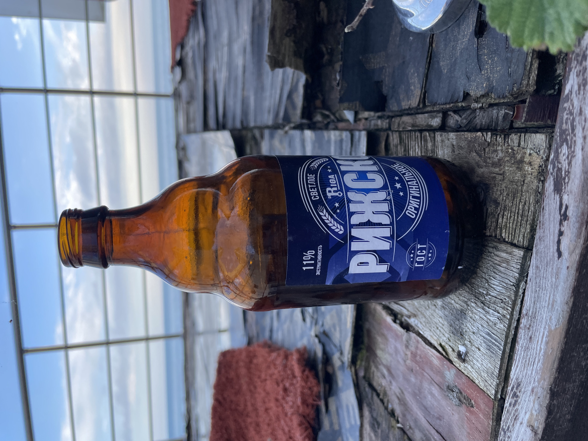A brown glass bottle of "Rizhskoe" beer lies on a weathered wooden surface. The bottle is partially filled, with the label prominently displayed. The label is blue and white, with Russian text and stars. The wooden surface appears to be part of a rustic structure, possibly a table or bench, and shows signs of age and wear. The background includes a window or glass structure, revealing an outdoor scene with a cloudy sky and potentially a body of water. The location is Olginka, Russia based on the beer brand.