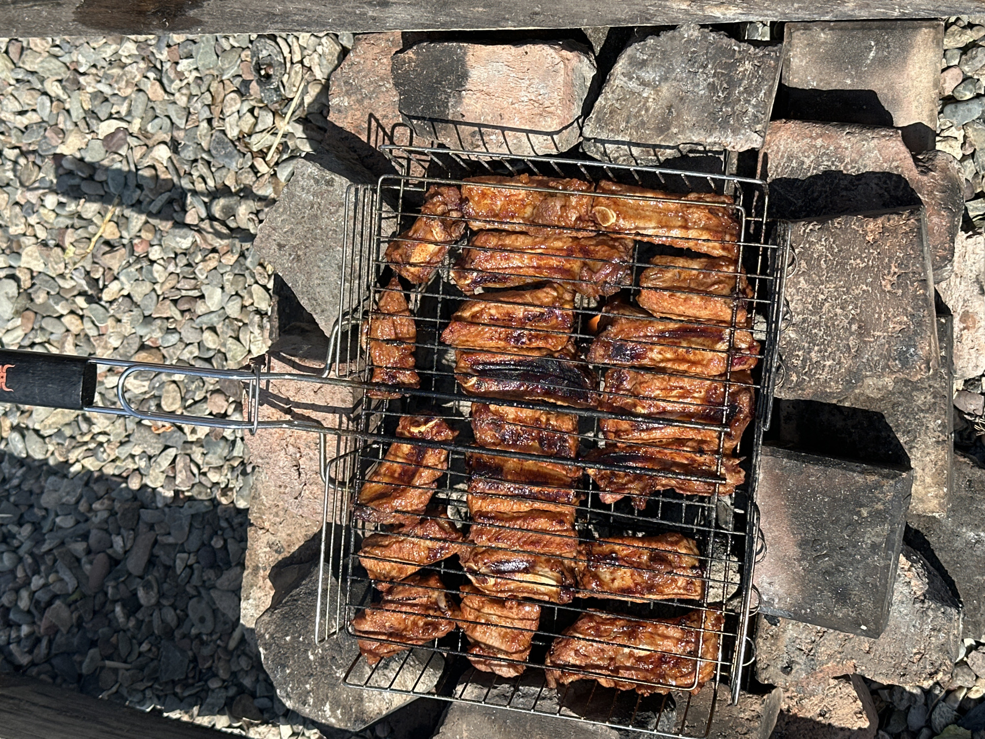 A metal grill basket filled with cooked ribs rests on a pyre of rocks. The ribs are a deep brown color, indicating they have been grilled to a well-done state. The grill basket itself is made of chrome-plated metal and has a handle at the top. The pyre is constructed from various sizes of rocks, with some larger, flat stones forming a base for the grill. The ground surrounding the pyre is covered in smaller gravel. The scene appears to be outdoors, with sunlight casting shadows on the ground and the rocks. The image has a rustic and informal feel, suggesting a casual outdoor cooking event. The location is not specified in the image, however, the prompt states it is in Novonikolaevka, Russia.