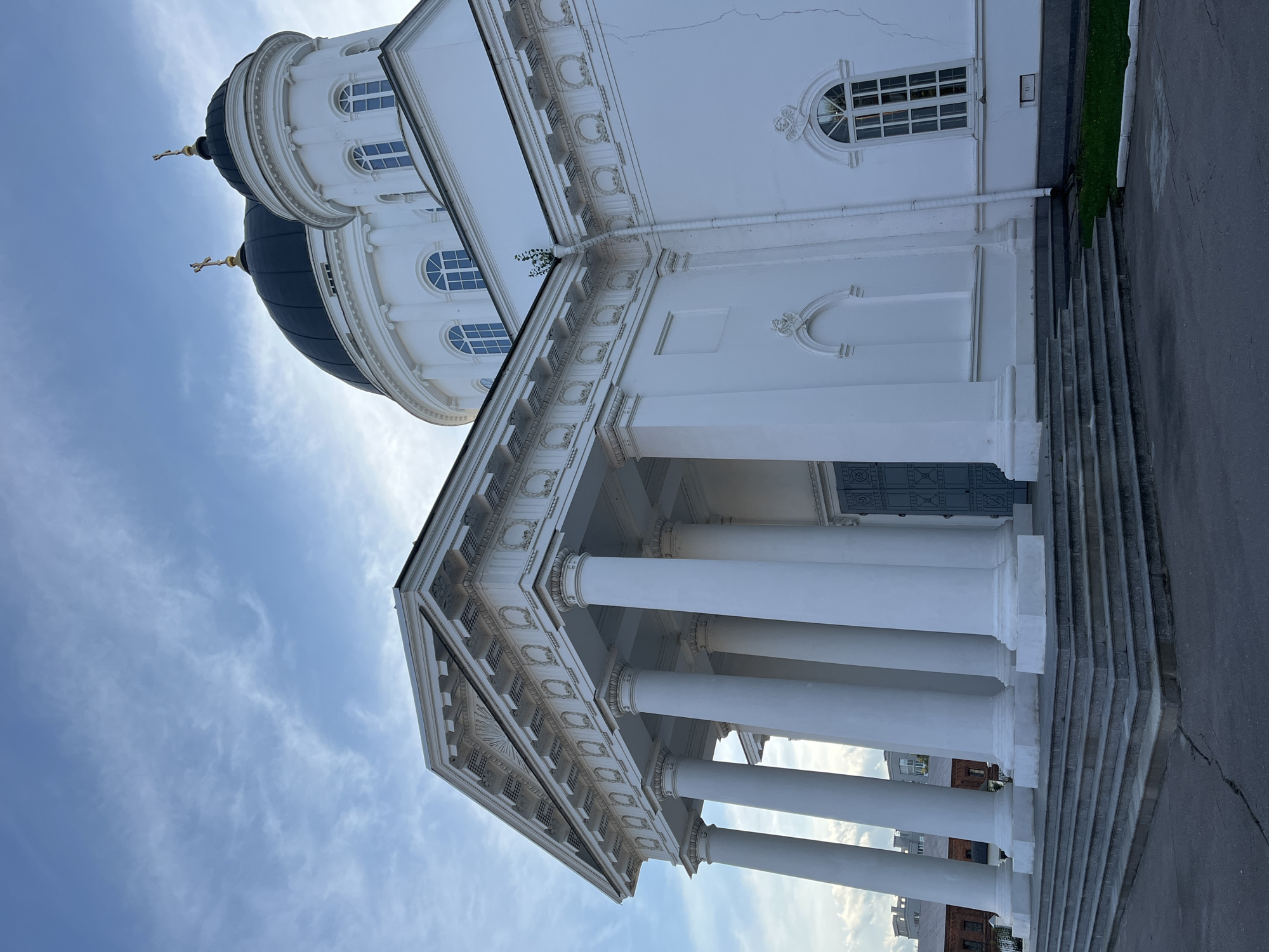 The image shows the exterior of a white neoclassical cathedral with large columns and domes, located in Nizhny Novgorod, Russia. The cathedral is a prominent architectural structure, likely a landmark. The sky above is blue with scattered clouds, suggesting it's either morning or late afternoon. There are no people or animals visible in the frame. The foreground shows a grey paved area with stairs leading up to the entrance of the building. The building itself appears well-maintained, with a grand and imposing presence.