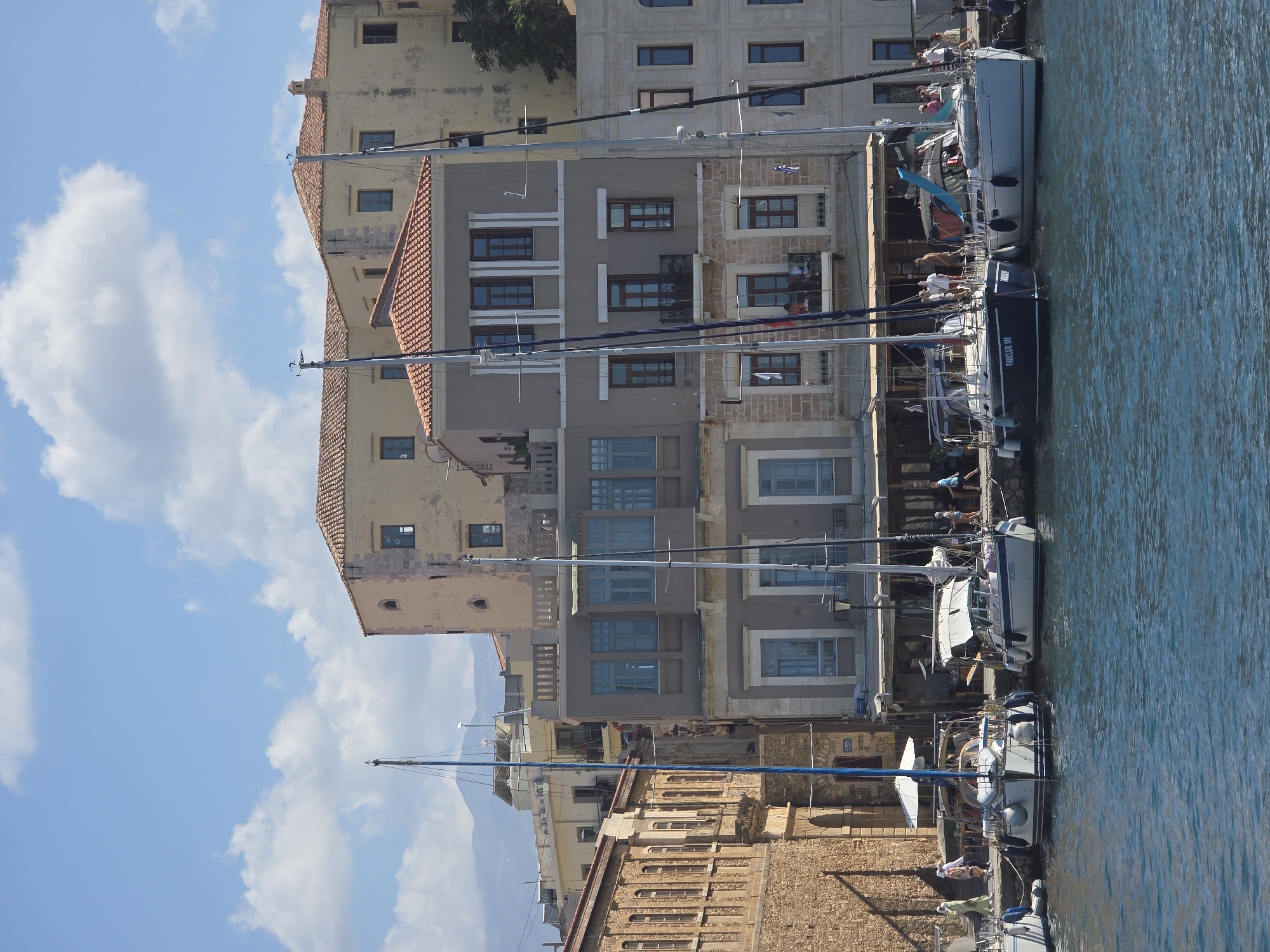 This is a photograph taken at the Venetian Harbor in Chania, Greece, on a sunny day with scattered clouds. The foreground features the deep blue water of the harbor. Moored along the stone quay are several sailboats. Two prominent sailboats are docked side-by-side; the left one is dark blue with "FAN CLUB VII" written on its side, and the right one is white. To the left of these, another white sailboat is visible, and a portion of yet another sailboat is seen on the far left edge of the frame.
Behind the sailboats, a row of buildings lines the harbor. The architecture is characteristic of Chania's Old Town, featuring stone facades, arched doorways, and weathered plaster in shades of beige, ochre, and gray. Many of these buildings have red-tiled roofs. Numerous windows, some with shutters, and balconies are visible.
Several people are present in the scene. On the quay, a group of individuals are walking or standing near the moored boats and the buildings. Some are dressed casually for a warm day. The overall impression is a picturesque harbor scene in a historic Mediterranean city.
