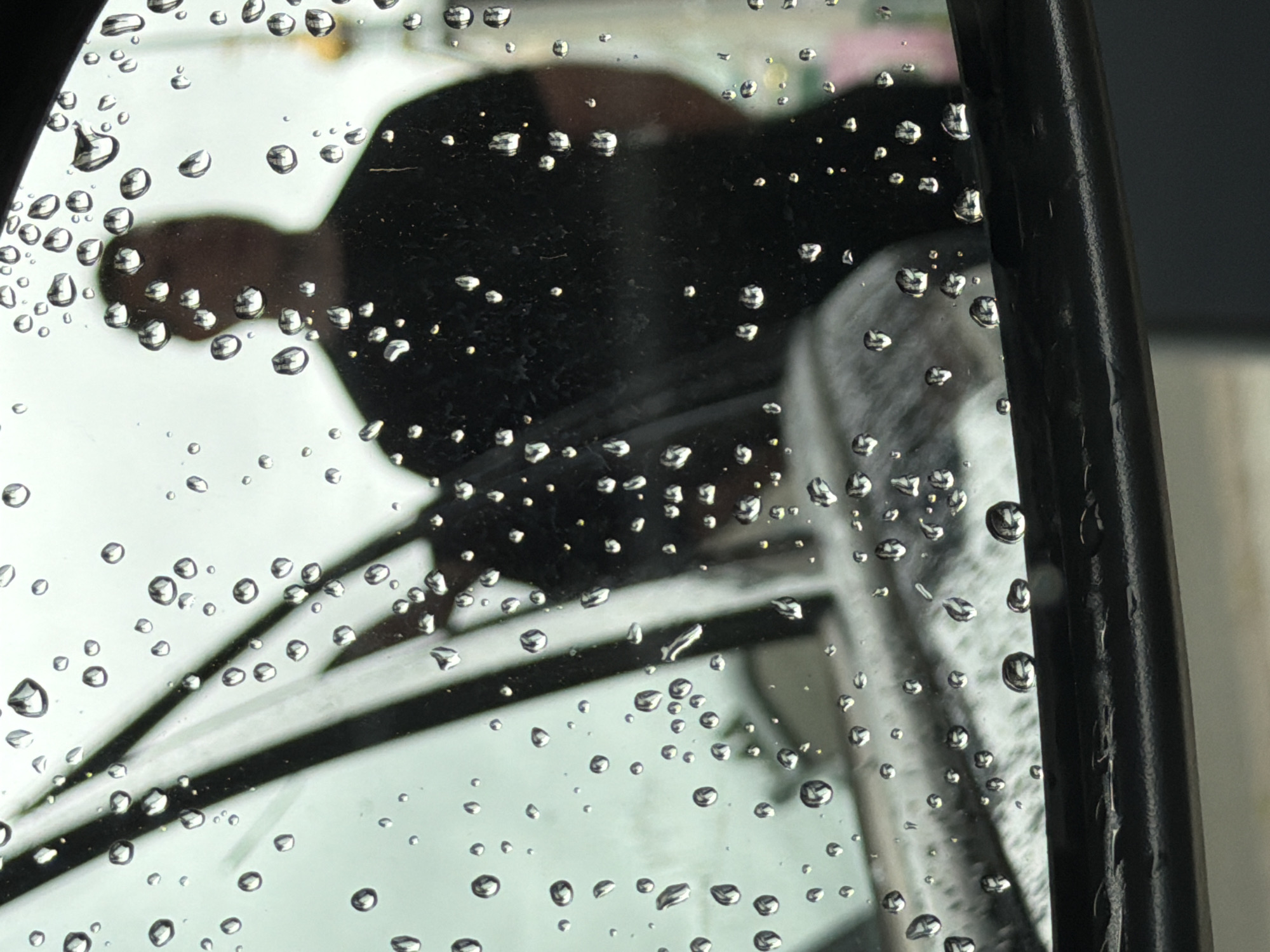 This image captures a close-up, distorted reflection in a rain-splattered car side mirror in the city of Chaikovsky, Russia. The mirror's surface is heavily covered with numerous clear water droplets of various sizes, creating a textured, refractive foreground. The black frame of the side mirror runs horizontally across the bottom edge of the image.
Reflected in the mirror is the dark, blurred silhouette of an individual, predominantly occupying the central and right portions of the frame. The person appears to be wearing a dark, possibly black, short-sleeved top, with their head and upper torso clearly outlined against a lighter background. The individual's face is obscured by the lack of detail in the silhouette and the overlay of raindrops. To the left of the person's silhouette, the reflection of a section of the car's interior or windshield frame, characterized by diagonal dark lines, is visible through the raindrops. The lower part of the mirror reflects the dark bodywork of the vehicle itself. The overarching background, visible through the reflections and raindrops, is a soft, light grey-green, suggesting an overcast sky or diffused light. Faint, indistinct yellow and white blurs are marginally visible in the far-right background. The scene suggests an outdoor environment during precipitation.