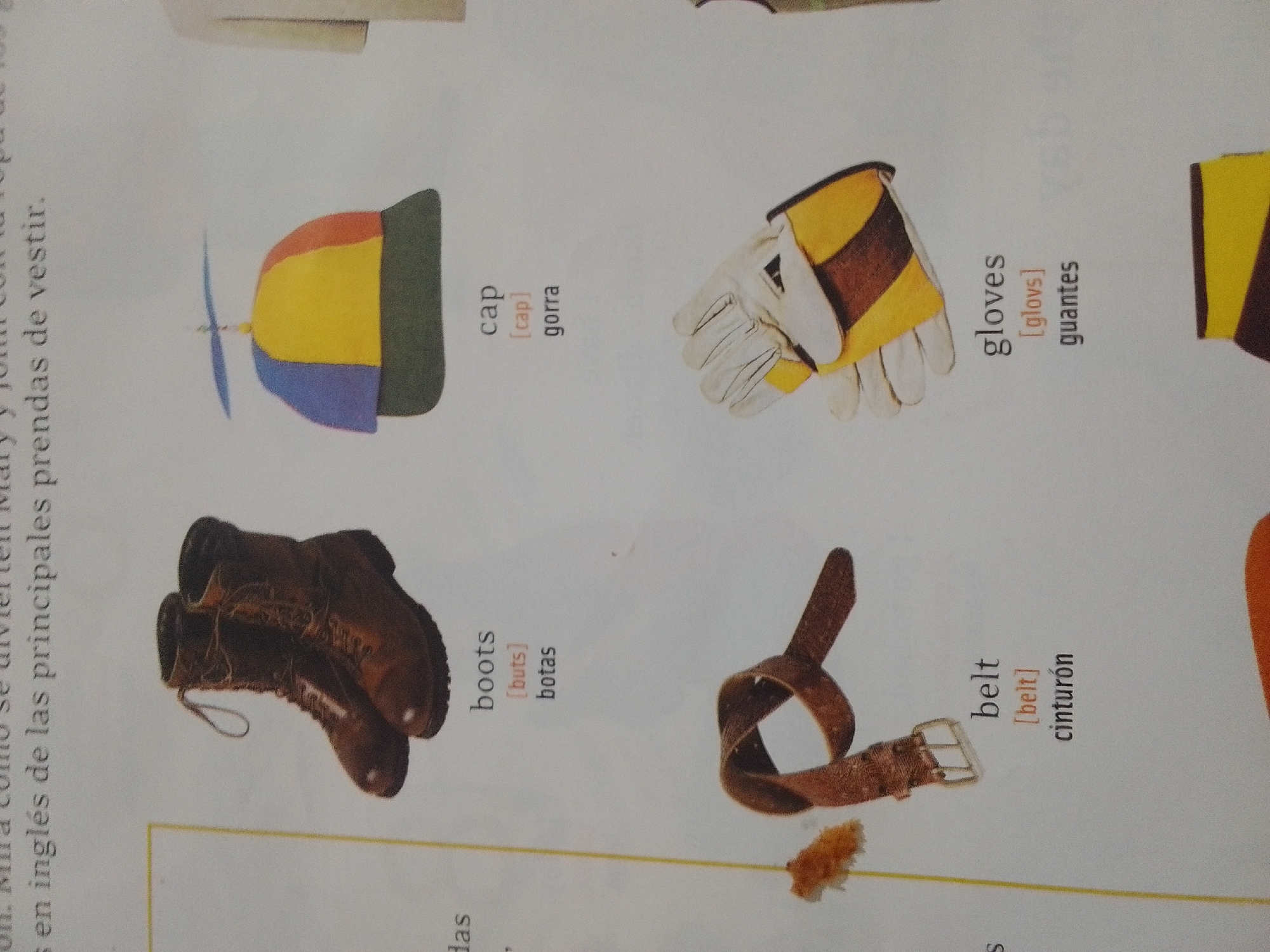 The image is a close-up, overhead shot of a page from a book or magazine. The page displays illustrations of various clothing items with their English and Spanish names. In the top left quadrant, there is a colorful cap with a propeller. To its right, there are a pair of white and yellow gloves with brown accents. Below the cap, there is a pair of brown leather boots. To the right of the boots, a brown leather belt with a buckle is shown. The text accompanying the illustrations appears to be educational, likely teaching vocabulary related to clothing. The page has a white background with some faint lines and possibly other images or text visible at the edges, indicating it is part of a larger spread. There is no discernible city, town, or environment depicted in the image.
