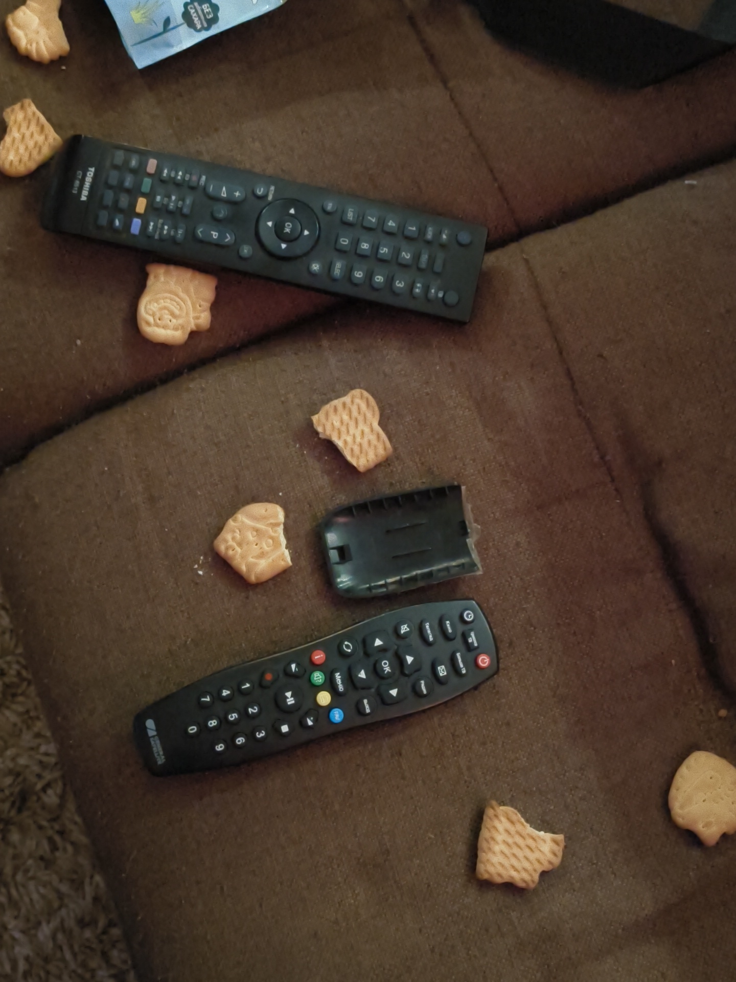 The image depicts a casual indoor scene, likely a living room in Chaikovsky, Russia, with two TV remotes and several cookies scattered across a brown, textured fabric surface, possibly a sofa. No people are visible.
A black "TOSHIBA CT-6513" remote control is positioned at the top of the frame. Its buttons include numbers 0-9, channel/volume controls, navigation arrows, and text such as "OK" and "LIST". Partially visible at the top left is a light blue and white package fragment bearing the Russian text "БЕЗ САХАРА" (Sugar-Free), suggesting the cookies' content.
The second, lower remote is also black and appears to be a universal or set-top box remote, with its battery cover detached and lying beside it. This remote also features number buttons (0-9), navigation, an "OK" button, and colored function buttons. Visible Russian text includes "Меню" (Menu), "Телегид" (TV Guide), "Больше ТВ" (More TV), and "Радио" (Radio).
Numerous small, light-colored cookies, some whole and others broken, are spread around the remotes. Their shapes vary, with one resembling an animal figure near the top remote and another with a waffle-like texture. The scattered items create an impression of a relaxed, possibly untidy, moment of snacking while watching television.