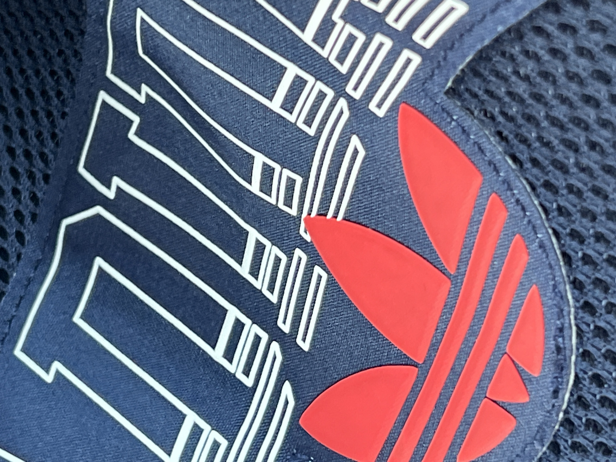 A close-up, high-angle shot shows a portion of a navy blue fabric item, likely a bag or clothing, featuring white outlined lettering and a red Adidas logo. The lettering appears to spell out "TEAM" with each letter formed by stacked rectangular shapes. The Adidas logo, a trefoil, is prominently displayed in bright red. The background is a dark, textured mesh fabric. The image does not provide any information about the city of Kinawley or the United Kingdom.