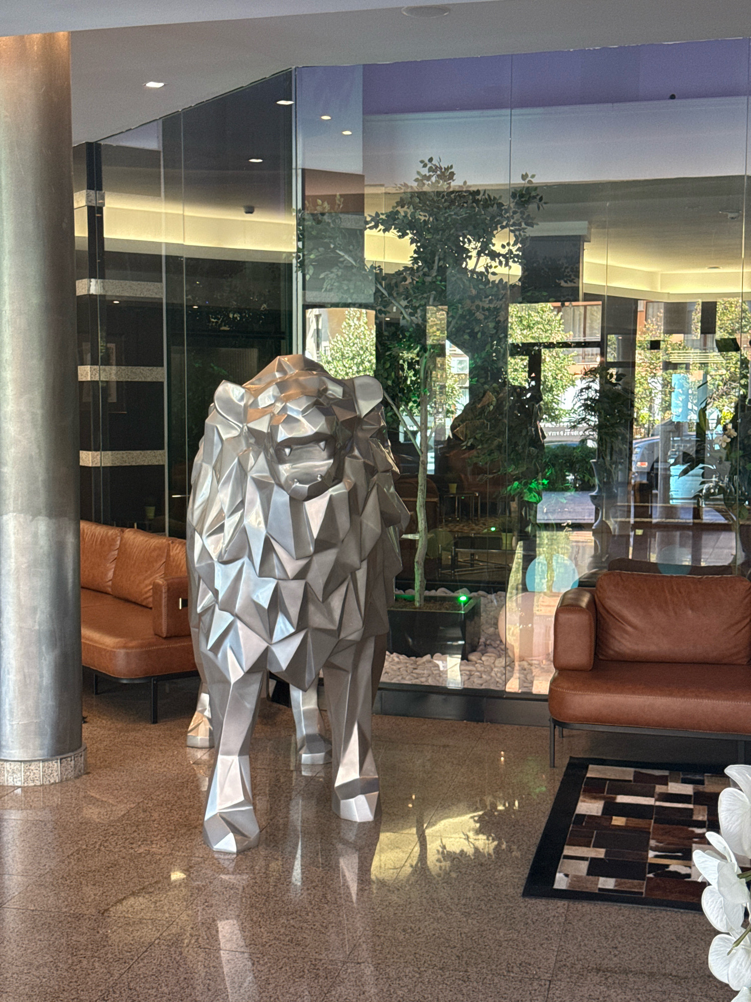 The image shows an interior scene, likely a hotel lobby or a modern office space. A large, geometric sculpture of a lion stands prominently in the foreground, made of a reflective, metallic material. To the left of the lion is a large, metallic cylindrical column. Behind the lion, a glass wall separates the interior from an outdoor or atrium area. Through the glass, one can see potted plants, trees, and reflections of the interior. Two brown leather sofas are visible, one to the left of the lion and partially obscured by the column, and another to the right. A small, patterned rug is placed in front of the sofa on the right. The flooring appears to be a speckled terrazzo. The lighting is a mix of ambient interior lights and natural light filtering through the glass. The image does not depict any people or animals. The provided context does not offer information about the location being Sveti Vlas, Bulgaria.