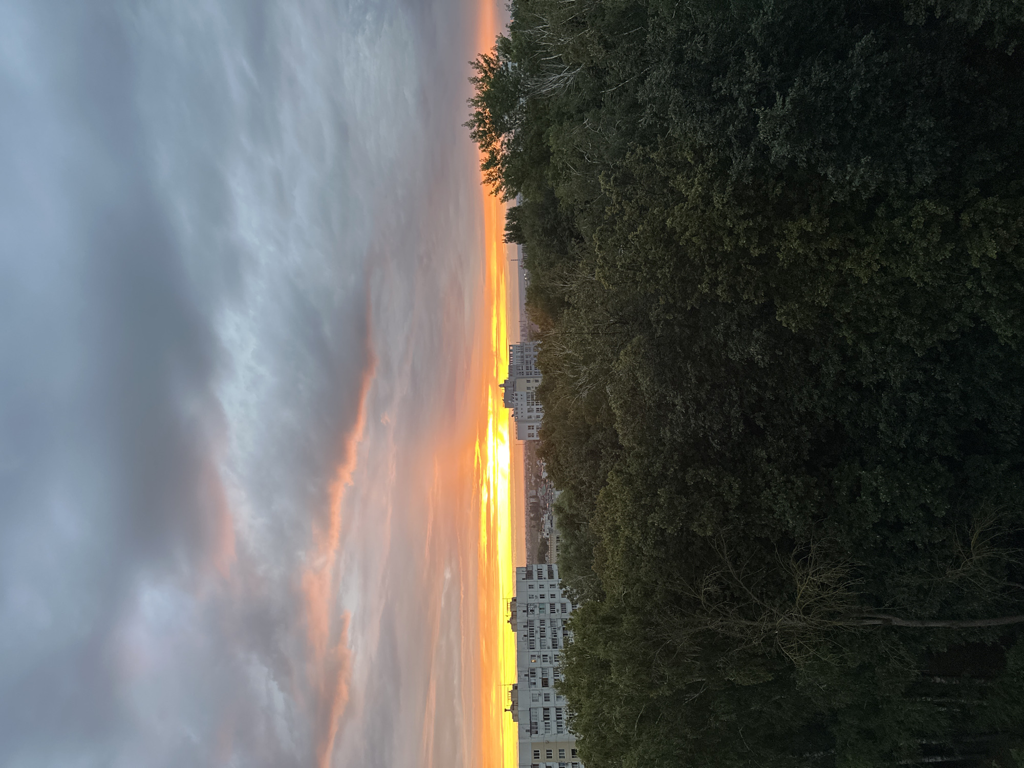 The image is a wide shot of a city at sunset, taken from a high vantage point. The sky is filled with dramatic clouds, tinged with orange and pink from the setting sun. The sun itself is a bright band of yellow on the horizon, with rays of light piercing through the clouds. In the foreground, there is a dense canopy of dark green trees. Behind the trees, on the left side of the image, are several multi-story apartment buildings. The buildings are white and have many windows. Further in the distance, more buildings and the outline of the city can be seen. The overall scene depicts an urban landscape at dusk, with the natural beauty of a sunset contrasting with the man-made structures. The location appears to be the city of Krugloye, Russia.