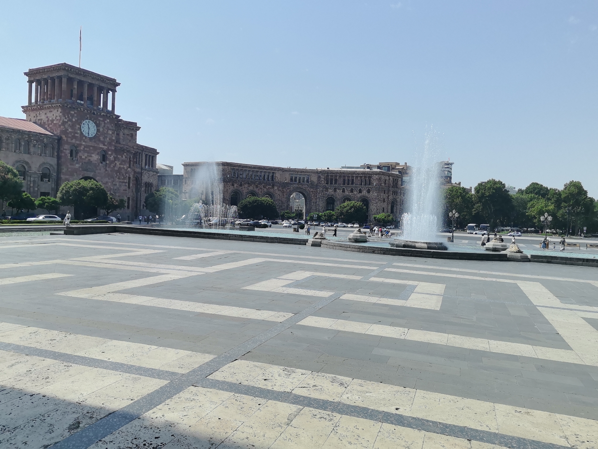 The image captures a wide-angle view of Republic Square in Yerevan, Armenia, on a clear, sunny day. In the foreground, a vast paved area is visible, featuring a geometric pattern of light and dark gray tiles, creating a striking visual design. Several fountains are active in the middle ground, with jets of water spraying upwards, catching the sunlight. Behind the fountains, a line of vehicles, including cars and buses, is parked or moving along a street.
Dominating the left side of the background is a grand, multi-story building with a classical architectural style. It features a prominent clock tower with Corinthian columns and a large circular clock face. Arched windows punctuate the facade of this building. To the right of this structure is another impressive building with a long, arcaded facade, characteristic of Armenian architecture. Trees with lush green foliage are interspersed between the buildings and behind the street, adding a touch of nature to the urban landscape.
Several people are scattered throughout the scene. Some are walking on the paved area, while others appear to be near the fountains or the buildings. The sky is a clear, light blue, indicating fair weather. The overall impression is of a well-maintained public space in a bustling city.