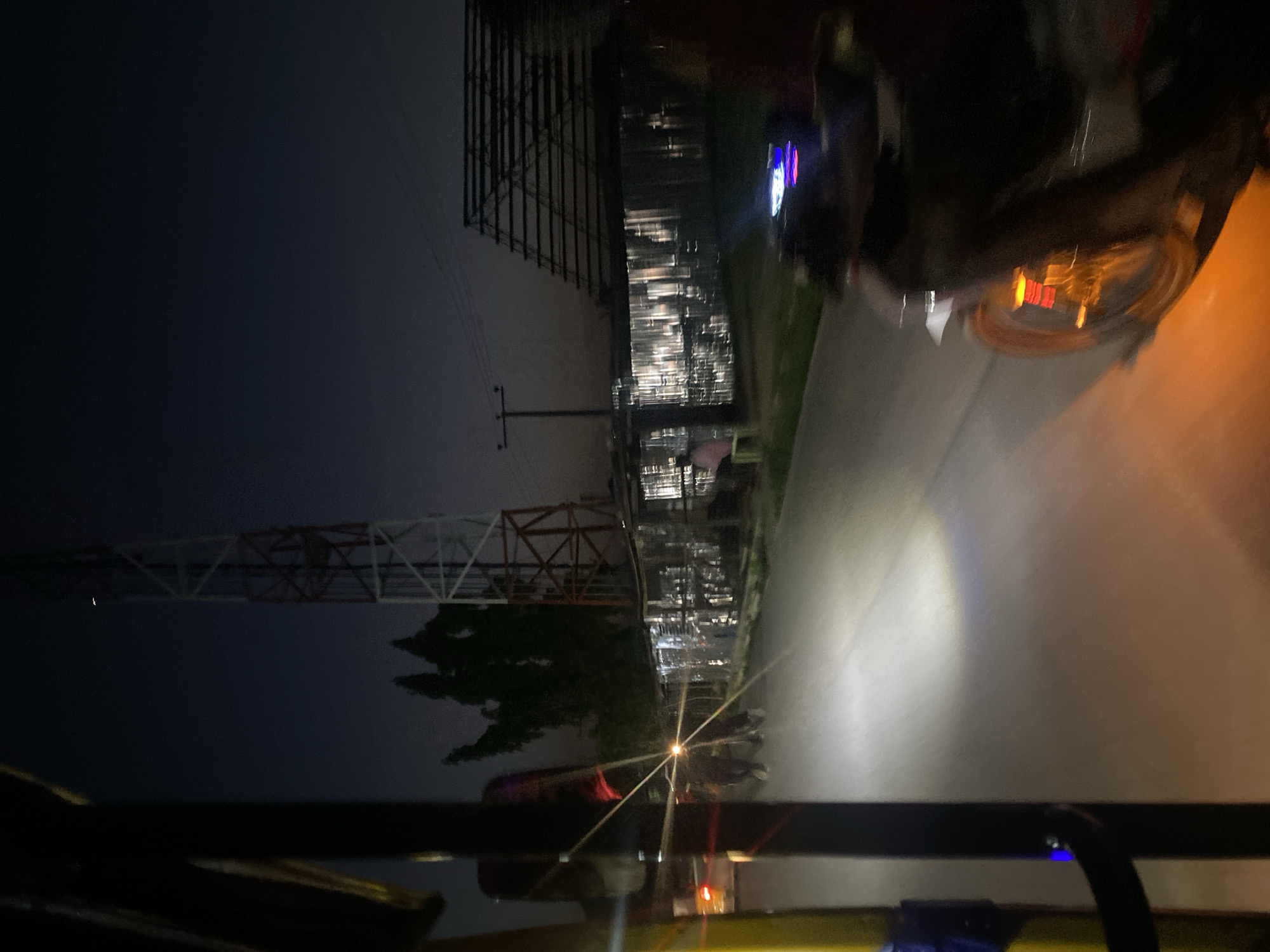 The media file shows a nighttime scene, likely in Jalingo, Nigeria, from the perspective of someone riding in a vehicle. The image is rotated, making it appear as if the scene is viewed sideways. In the foreground, the edge of a vehicle's interior is visible, including part of a dashboard with some lights.
The main subjects are the environment and infrastructure illuminated by artificial light. A large, lattice tower, possibly a transmission tower, stands prominently on the left side of the frame, against a dark, twilight sky. Further in the background, there are various structures that appear to be part of an industrial complex or a city skyline, with numerous lights glowing. The road is visible on the right, with a bright headlight beam cutting through the darkness. Several vehicles, possibly motorcycles, are clustered together on the road.
The time of day is clearly night, indicated by the darkness and the artificial lighting. There are no immediate cues about the weather, but the general darkness suggests it's not raining heavily. The presence of the transmission tower and the industrial-looking structures provide some location context, hinting at an urban or semi-industrial area. The overall mood is somewhat dynamic due to the motion implied by the perspective and the light trails. No legible text is visible in the image.