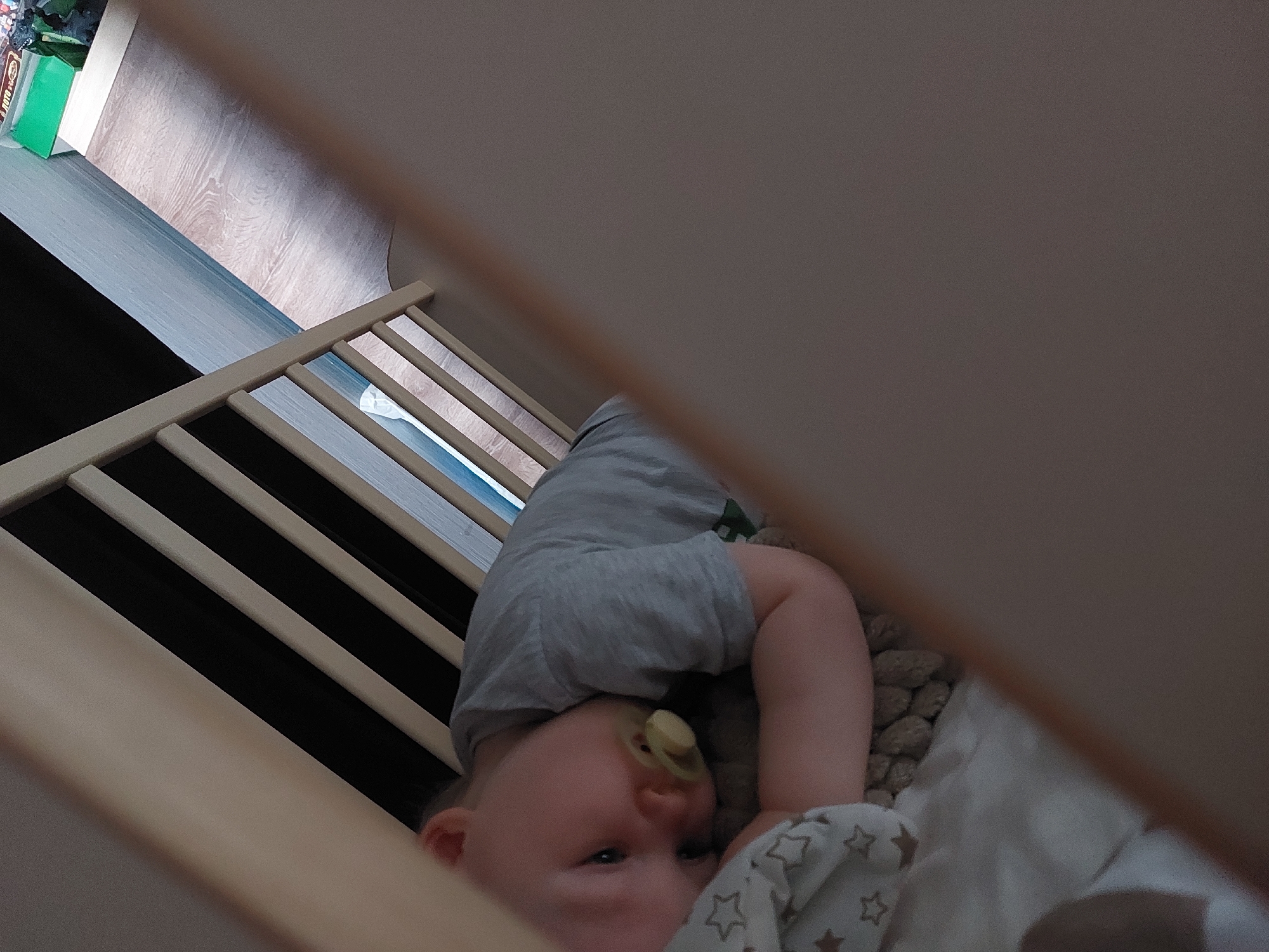 A baby wearing a gray shirt is lying in a crib with a pacifier in its mouth. A patterned blanket is partially covering the baby. The crib has wooden slats on one side. The floor and a box are visible in the background. The location is Chaikovsky, Russia.