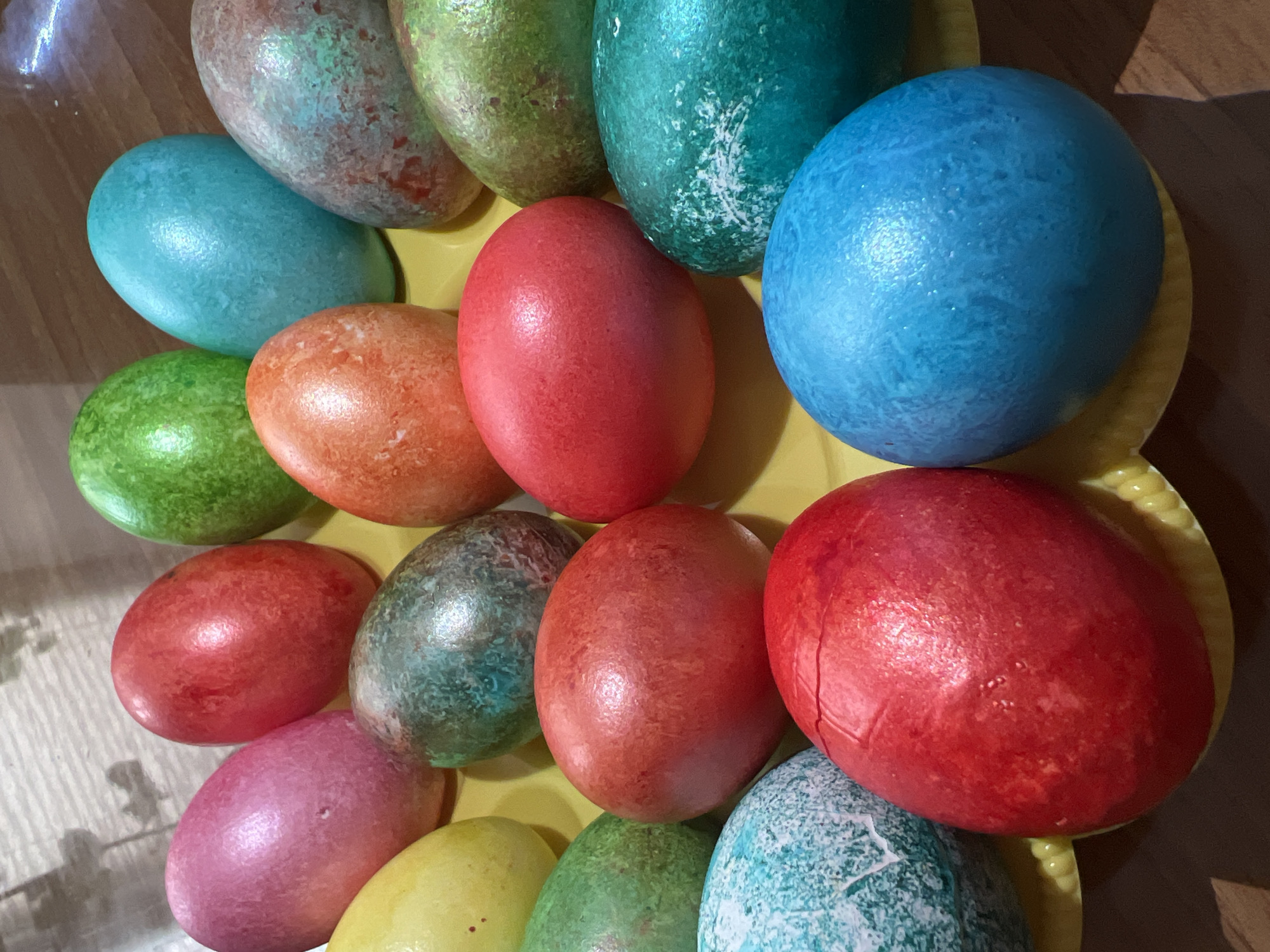The image depicts a collection of approximately 17 dyed Easter eggs of various colors including blue, green, red, orange, pink, and yellow arranged on a yellow plate. Some eggs show mottled or speckled patterns, likely from the dyeing process. The lighting appears to be artificial, creating shadows and highlights on the eggs. The scene is situated indoors. No information suggests this image to be connected to Votkinsk, Russia.