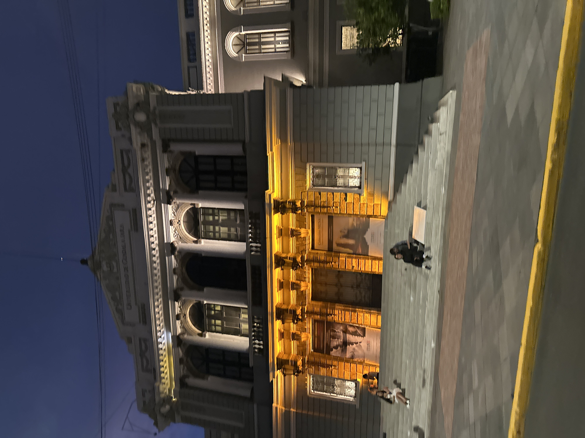 The image shows the University of Guadalajara's Paraninfo building at night. The building is illuminated with warm light, highlighting its neoclassical architectural features. A set of stairs leads to the building's entrance, where people are present. The sky is a deep blue, indicating it's late evening or night. The sidewalk is paved with gray bricks. A yellow curb separates the sidewalk from the street.