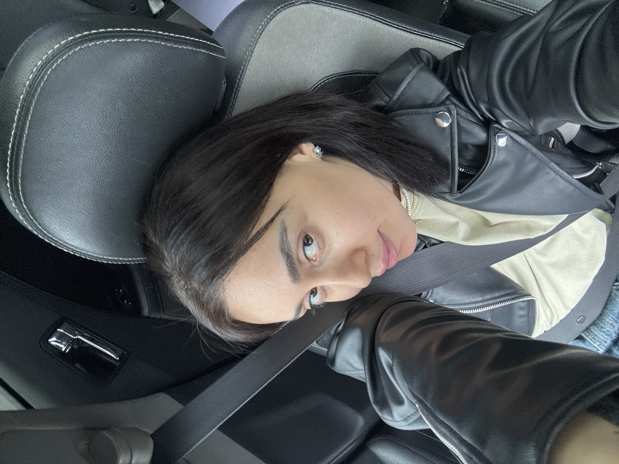 A female individual is depicted seated in the driver's seat of a vehicle. She has dark hair and brown eyes, looking directly at the camera. She wears a black leather jacket with silver-toned zippers and snaps, a light-colored top, and denim trousers. A grey seatbelt is fastened across her chest. Small light blue stud earrings are visible. The vehicle's interior features a black and grey upholstered seat with white stitching and a black door panel with a chrome handle. The specific location within Tbilisi, Georgia, is not discernible from the image.