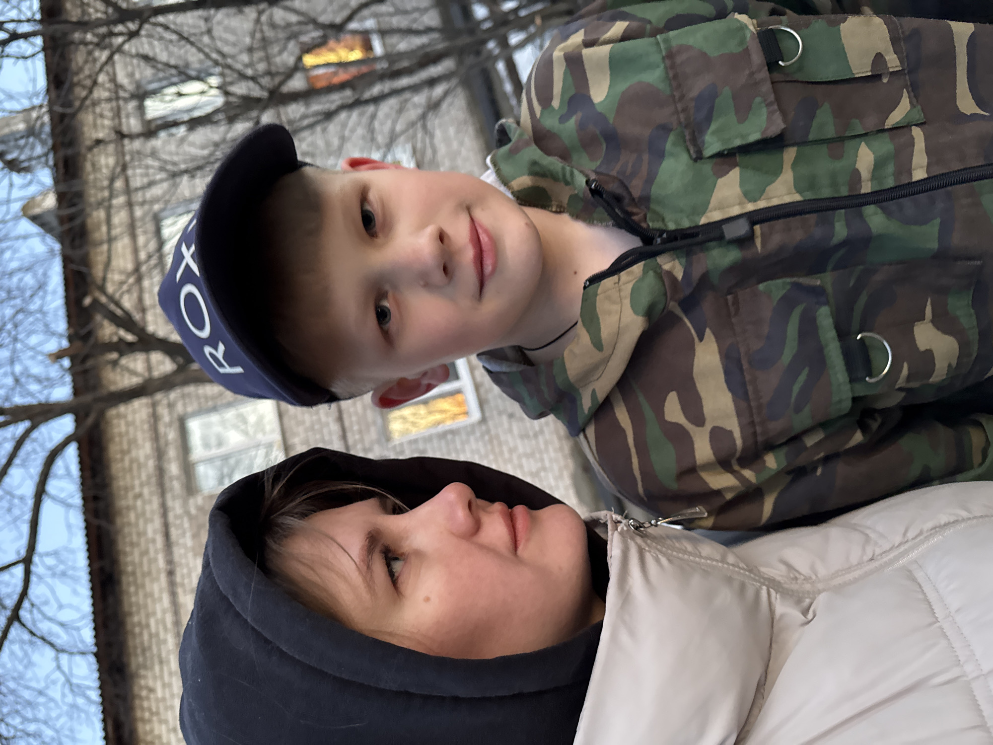 In Горный, Russia, a medium shot presents a woman and a boy posing together outside. The woman, wearing a light-colored puffer jacket with a dark hood up, looks up and to the left. A young boy stands above her and slightly to her right. He is wearing a camouflage patterned jacket and a dark baseball cap with white lettering. Bare branches and a building with windows are visible in the background. The sky is overcast.