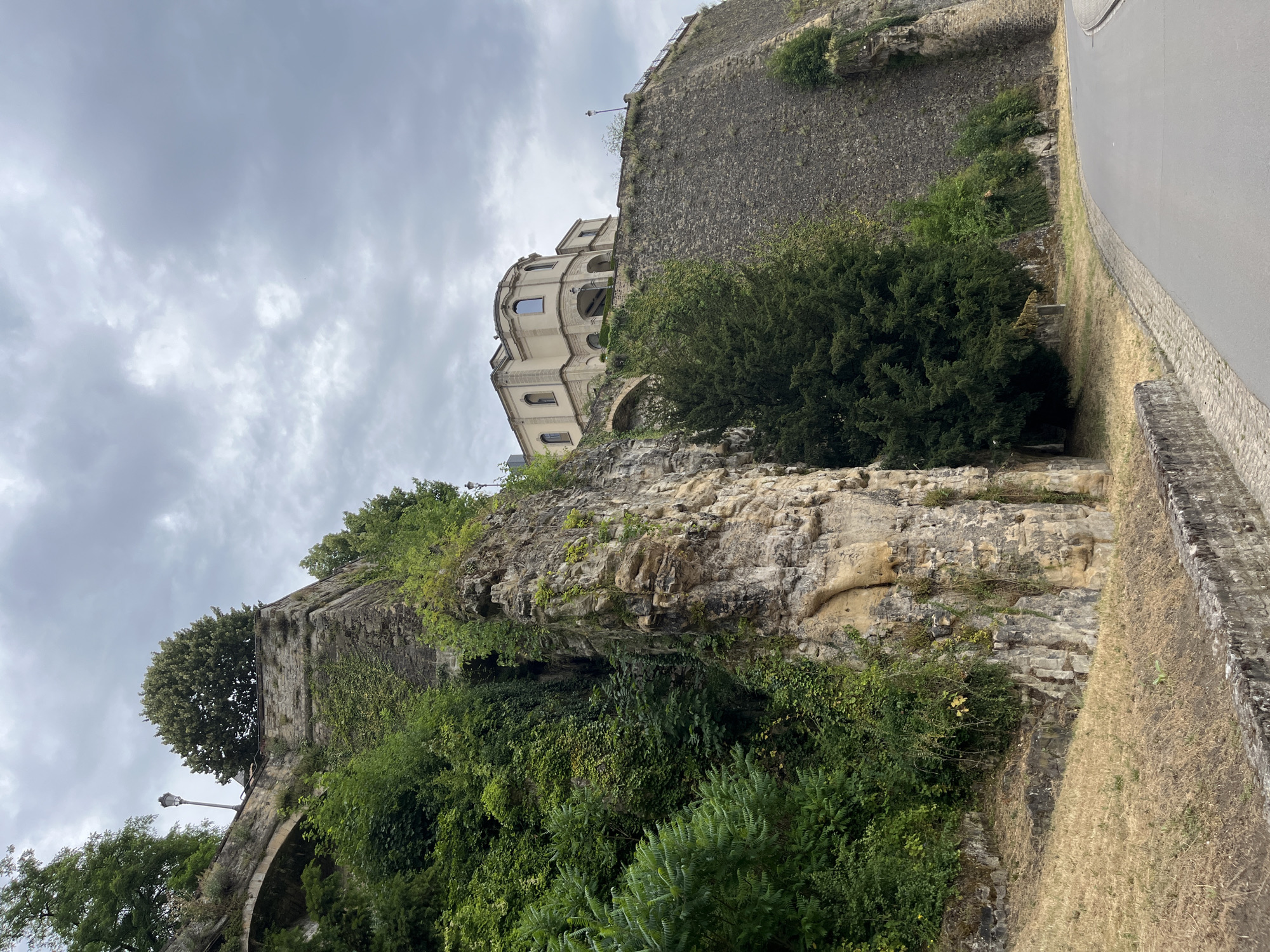This image captures a dramatic scene of a stone building perched atop a rugged, rocky cliffside, overgrown with lush green vegetation. The building, with its classical architecture featuring arched windows and balconies, appears to be a historic structure, possibly a fortress or a grand residence. The cliff itself is a natural formation of layered rock, further softened by climbing vines and dense foliage.
To the right of the scene, a paved road curves away, bordered by a stone retaining wall and dry, sun-bleached grass, suggesting a late summer or early autumn setting. The sky above is overcast with heavy, grey clouds, creating a subdued and somewhat dramatic atmosphere. There is no visible text in the image. The scene seems to depict a static, architectural and natural landscape rather than an active event. The overall impression is one of historical grandeur and natural resilience, with the building standing as a testament to human construction against the raw power of the earth.