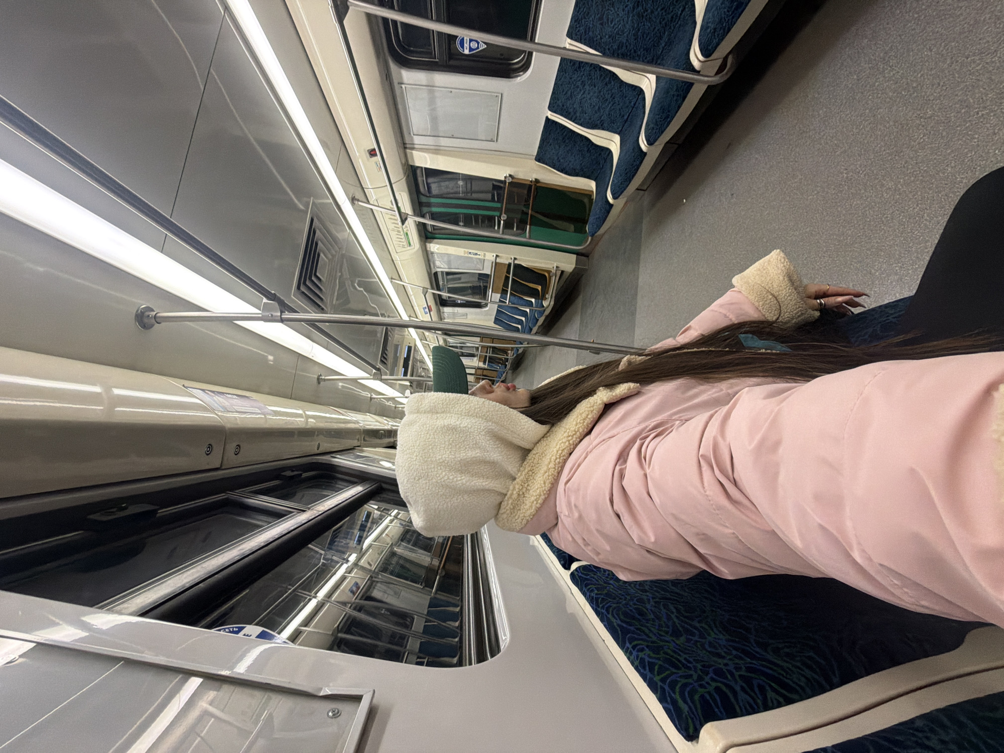 The image shows a person sleeping on a subway train. The person is wearing a pink coat and white hood. They are lying on the seat, using the window as a headrest. The subway car has blue and white seating. The scene is on the Saint Petersburg subway, Russia.