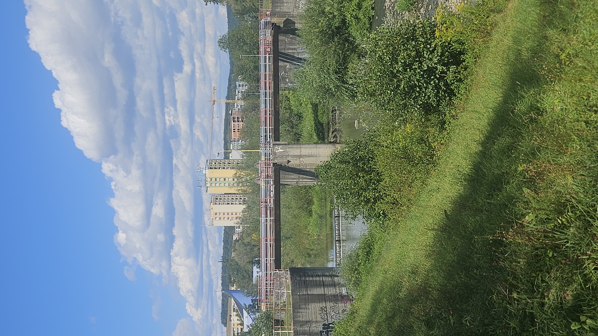 The image shows a view of a bridge over a river in the city of Prešov, Slovakia. The bridge is made of concrete and has metal railings. There are buildings in the background, including tall apartment buildings and a construction crane. The river is surrounded by lush green vegetation. The sky is blue with some white clouds. The scene appears to be a sunny day.