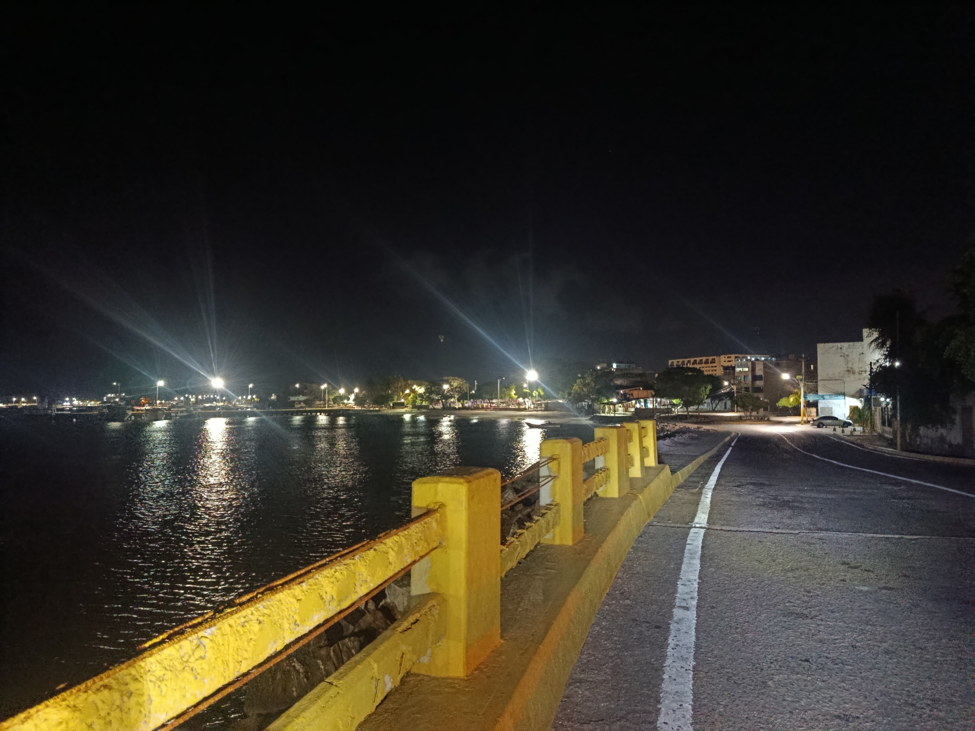 The image depicts a night scene in Juan Griego, Venezuela. A road runs alongside a body of water, possibly a bay or ocean. A yellow-painted barrier separates the road from the water. Streetlights illuminate the scene, creating reflections on the water's surface. In the distance, buildings and infrastructure are visible, defining the urban coastline. A white line marks the edge of the road. A car is parked further down the street. The sky is dark, with some light emanating from the urban area.