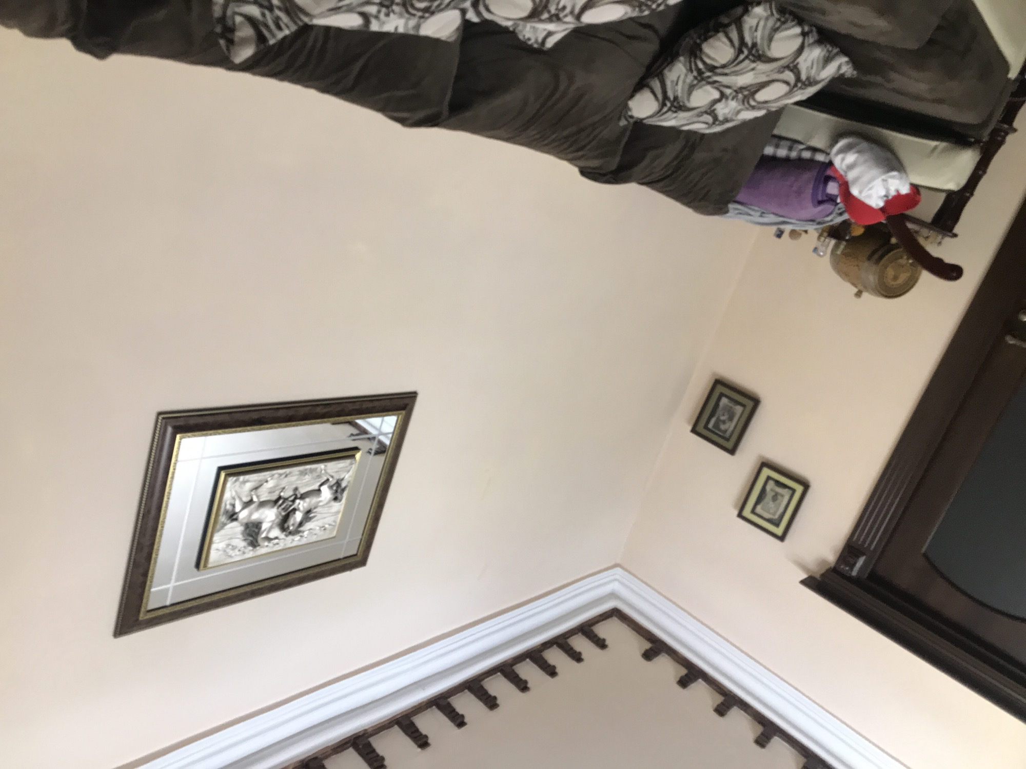 The image depicts an interior scene, possibly a living room, with a couch partially visible in the upper section of the frame. The couch is upholstered in a dark brown material and adorned with patterned pillows. Below the couch, on the right side, a barrel-shaped object with possible decorative elements is observed, along with a stuffed animal or toy with red and white coloring. The walls are painted in a light peach or beige hue. On the wall, several picture frames are hanging. A large, diamond-shaped frame featuring an image of galloping horses is prominent on the left wall. Further to the right, two smaller, square frames with dark borders are hung on the wall closer to a dark-colored doorway or window frame. In the lower section of the image, a decorative molding or cornice is present, suggesting architectural details. The scene's location, Yerevan, Armenia, is not discernible from the image alone.