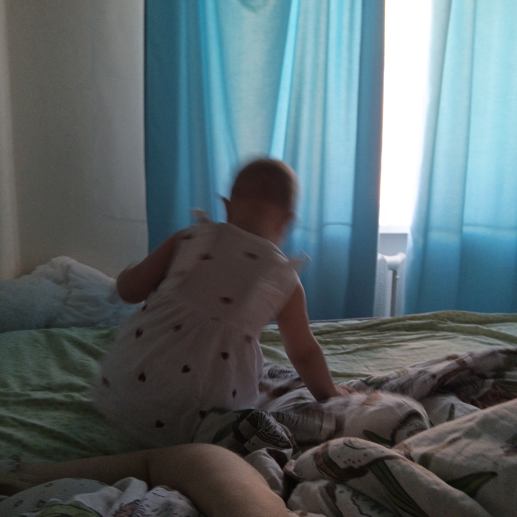The photograph shows the back view of a child in a white dress with small red heart prints, standing on a bed with green and patterned bedding. The child's head is slightly blurred, suggesting movement. A person's arm is partially visible on the bed. In the background, blue curtains frame a window with bright light shining through. The location is an interior bedroom space. No city information is available.