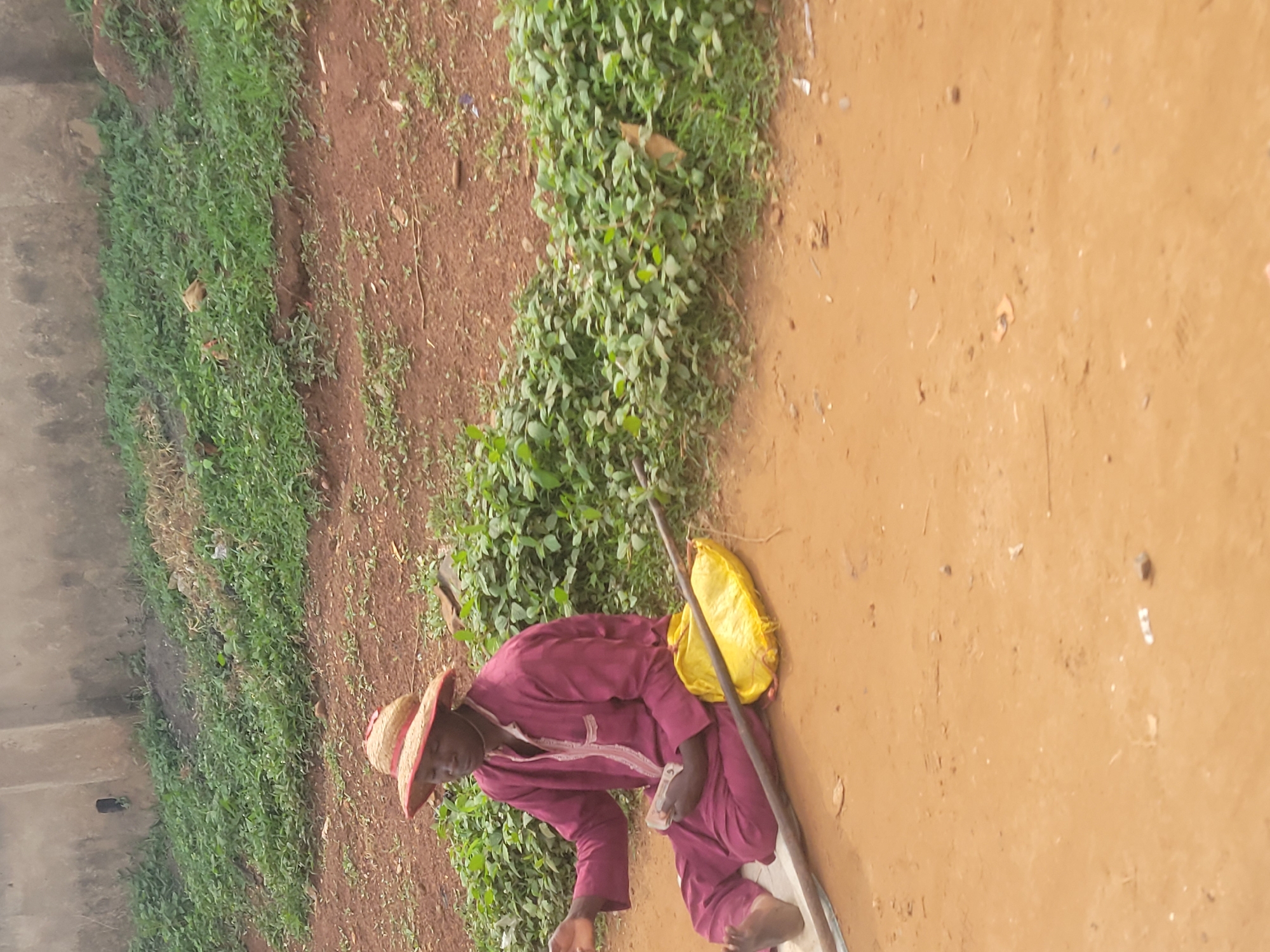 A person wearing a straw hat and a maroon robe sits on the ground next to a yellow bag. The person is holding a wad of cash in their right hand and appears to be counting it. A stick lies on the ground in front of the person, and a wall is visible in the background. The scene is outdoors in a dusty, unpaved area with some vegetation. The location is the city of Keffi, Nigeria.