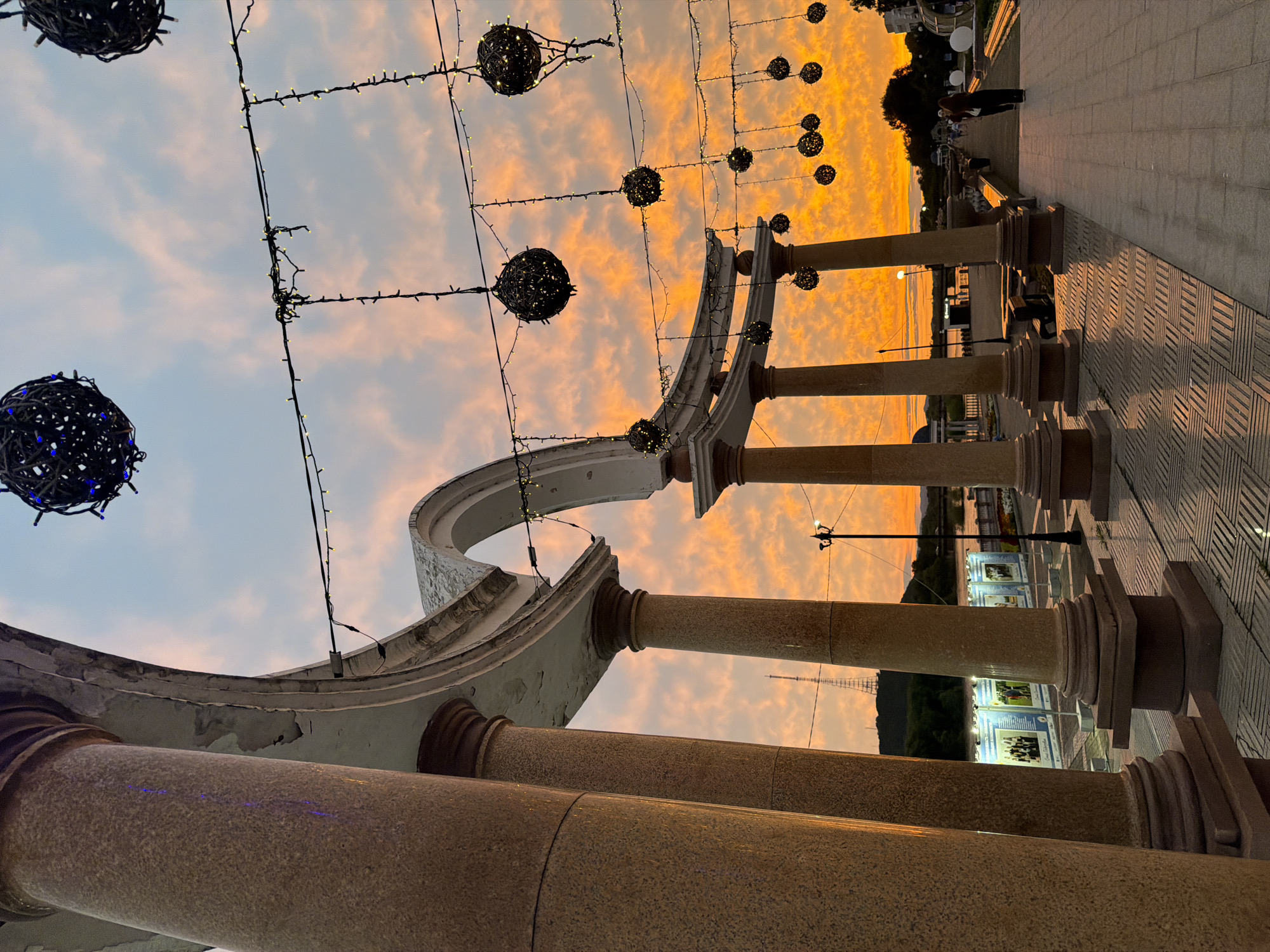 This is a low-angle, wide shot of a waterfront promenade in Birobidzhan, Russia, at sunset. The sky is ablaze with vibrant orange and pink hues, indicating the sun is low on the horizon. In the foreground, large, granite-like columns with ornate bases support an arched structure. String lights with decorative woven spheres are strung across the promenade, some with blue and yellow bulbs. Beyond the columns, a paved walkway stretches into the distance, lined with streetlights and a few people walking. A river is visible to the left, with a transmission tower and a hill in the background. The promenade appears to be an area for public gathering, possibly a park or esplanade, with posters displayed on stands near the riverbank. The overall atmosphere is serene and picturesque, characteristic of a summer evening in a Russian city.