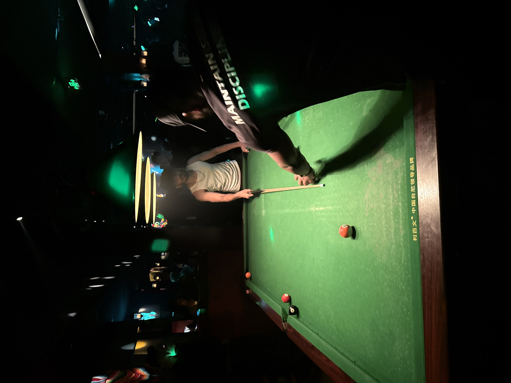 A dimly lit pool hall in Liangjiang, China, features two men engaged in a game of billiards. The man in the foreground, wearing a black t-shirt with the words "MAINTAIN SELF DISCIPLINE" printed in reflective green lettering, is positioned behind the pool table, cueing a shot. A second man, wearing a white tank top, stands across the table, observing the game. The green felt of the pool table is visible, with several billiard balls scattered across its surface, including a red ball, a black ball, and an orange and white striped ball. The environment is characterized by low lighting, with green spotlights casting an atmospheric glow over the scene. In the background, other patrons and decorative elements of the pool hall are partially obscured by the darkness.