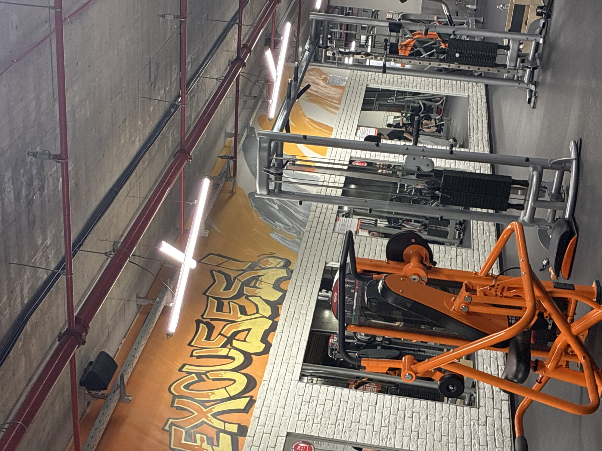 A gym in Plovdiv, Bulgaria, features fitness equipment arranged in rows along a wall adorned with a mural of the word "EXCUSE," painted in orange and yellow. Red pipes run horizontally along the concrete ceiling. Rectangular fluorescent lights are mounted to the wall. Exercise machines with weight stacks are positioned adjacent to white brick walls with mirrored panels. People are using the equipment, visible in the mirrors. The gym floor is gray. The angle of the shot is angled upwards.