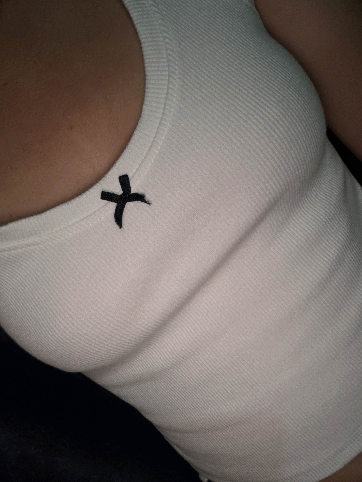 The image shows a close-up of a person wearing a white ribbed tank top with a small black bow on the left side of the neckline. The tank top is form-fitting and highlights the contours of the wearer's chest. The skin tone appears to be light brown. The background is dark and out of focus. There is no identifiable location in the city of Phan Thiết, Vietnam, or any environmental context. The image is a still life focusing on the garment and the part of the body wearing it.