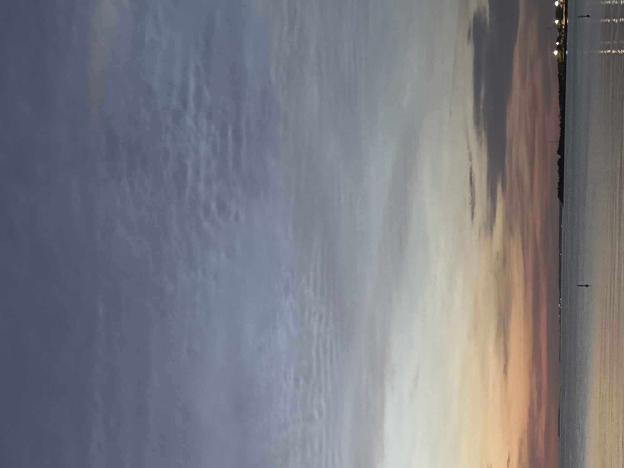 The image depicts a serene seascape at dusk or dawn, with a vast expanse of sky meeting the calm water. The sky transitions from a soft orange and pink hue near the horizon, characteristic of twilight, to a muted blue-gray higher up. Wispy clouds are scattered across the sky, some appearing as delicate streaks, others as larger, more diffused formations. The water's surface is largely smooth, reflecting the subtle colors of the sky.
In the distance, a faint, dark landmass is visible, dotted with what appear to be distant lights, suggesting a coastal town or city. There are a few darker shapes on the water, possibly buoys or small boats, and a single, slender vertical object, likely a marker or a small buoy, is present in the foreground water. The overall atmosphere is peaceful and tranquil.
While the image provides a general view of a coastal environment during twilight, it does not contain any specific landmarks or details that would definitively place it within the city of Tallinn, Estonia.