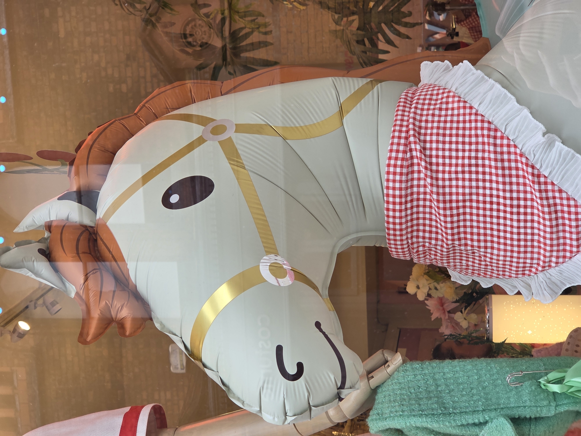 The image shows a horse balloon dressed in a red and white checkered dress with a white frilly edge. The balloon is primarily light green with brown mane and tail. It features gold trim around its face. The horse's eyes are brown, and it has a simple black smile. The balloon appears to be part of a shop window display, as reflected images of a building and foliage can be seen on the balloon's surface. The exact location is the city of Unknown, Unknown.