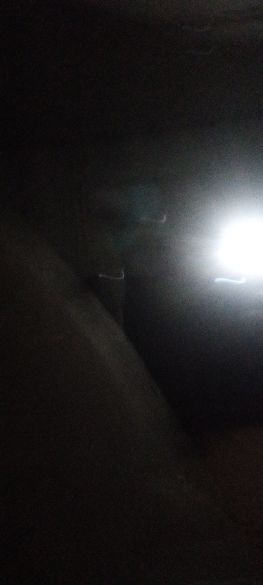The image is very dark and blurred. There is a bright light source in the upper right portion of the frame. In the lower left, there appears to be a dark, solid surface. The location appears to be inside. The city is unknown.