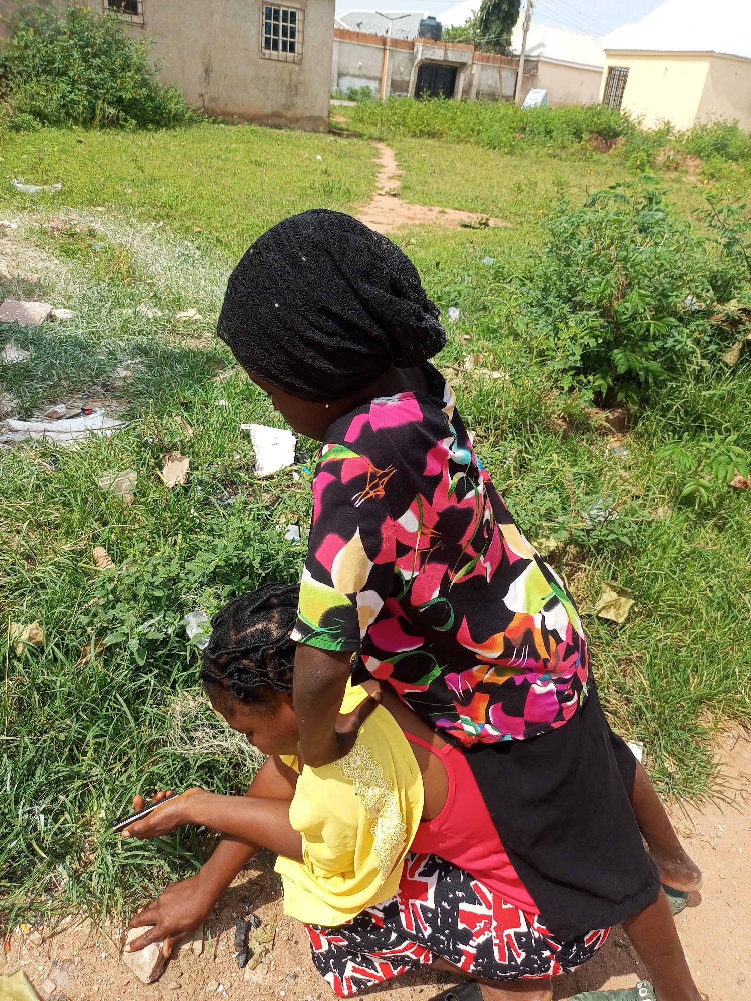 Two young girls are in a grassy outdoor area with some dirt patches. One girl is seated and leaning forward, holding a pen and a rock in her hands. She is wearing a yellow top and a patterned skirt. The other girl is standing and leaning over the seated girl, her body partially obscuring the seated girl. She is wearing a black headscarf and a colorful, abstract patterned shirt. The background shows a building with windows and some greenery. The scene appears to be in Tempure, Nigeria, on a bright, possibly sunny day. The activity seems to be one girl showing or teaching the other something with the pen and rock, possibly drawing or writing on the ground.