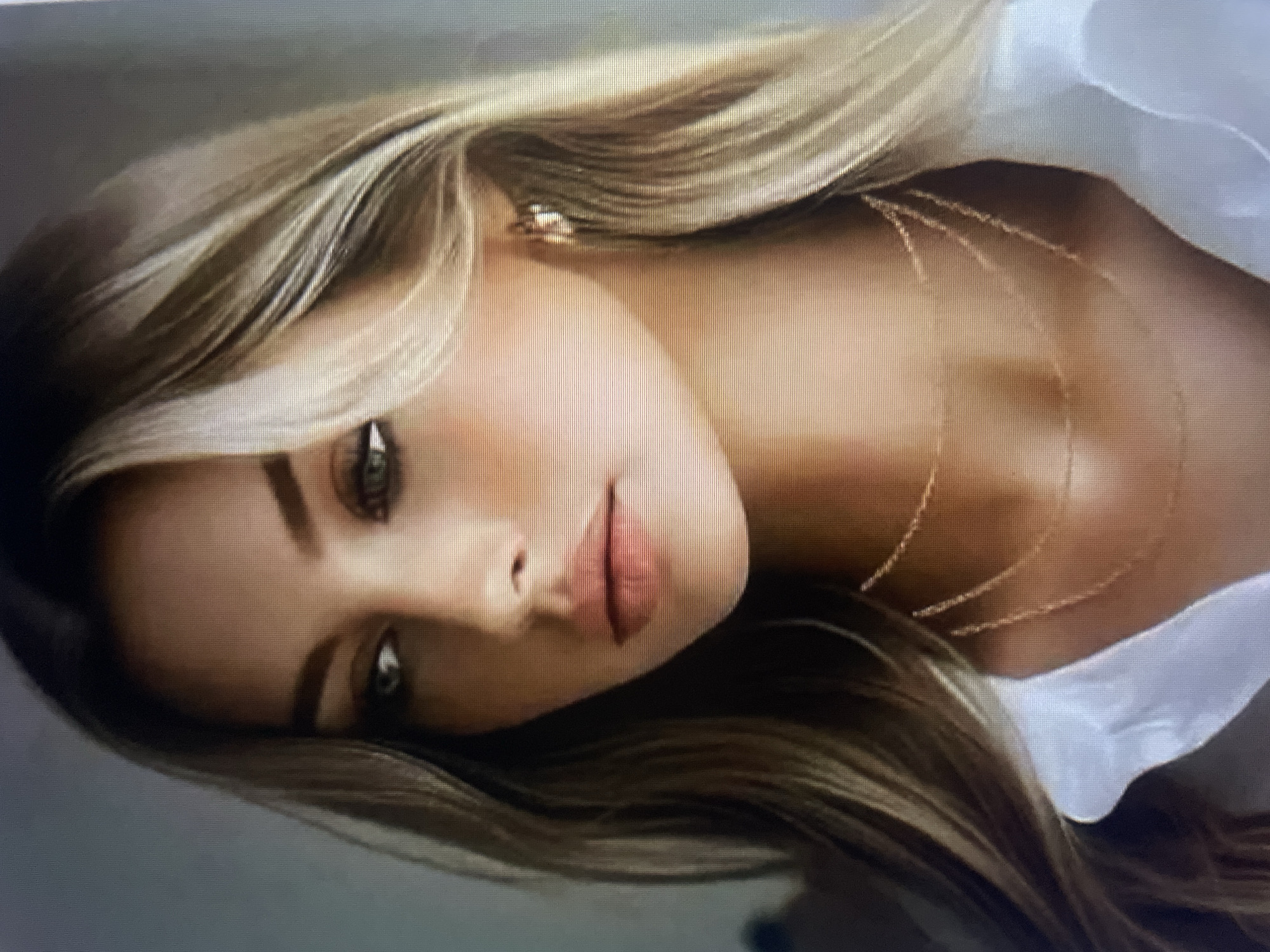 This is a close-up, waist-up studio portrait of a young woman with long, wavy, blonde and brown highlighted hair. She is wearing a white collared shirt and a multi-strand gold necklace. She has striking green eyes, prominent cheekbones, and full lips. A small gold earring is visible on her right ear. The background is a soft, out-of-focus light color. There is no discernible location in the city of Kołobrzeg, Poland.