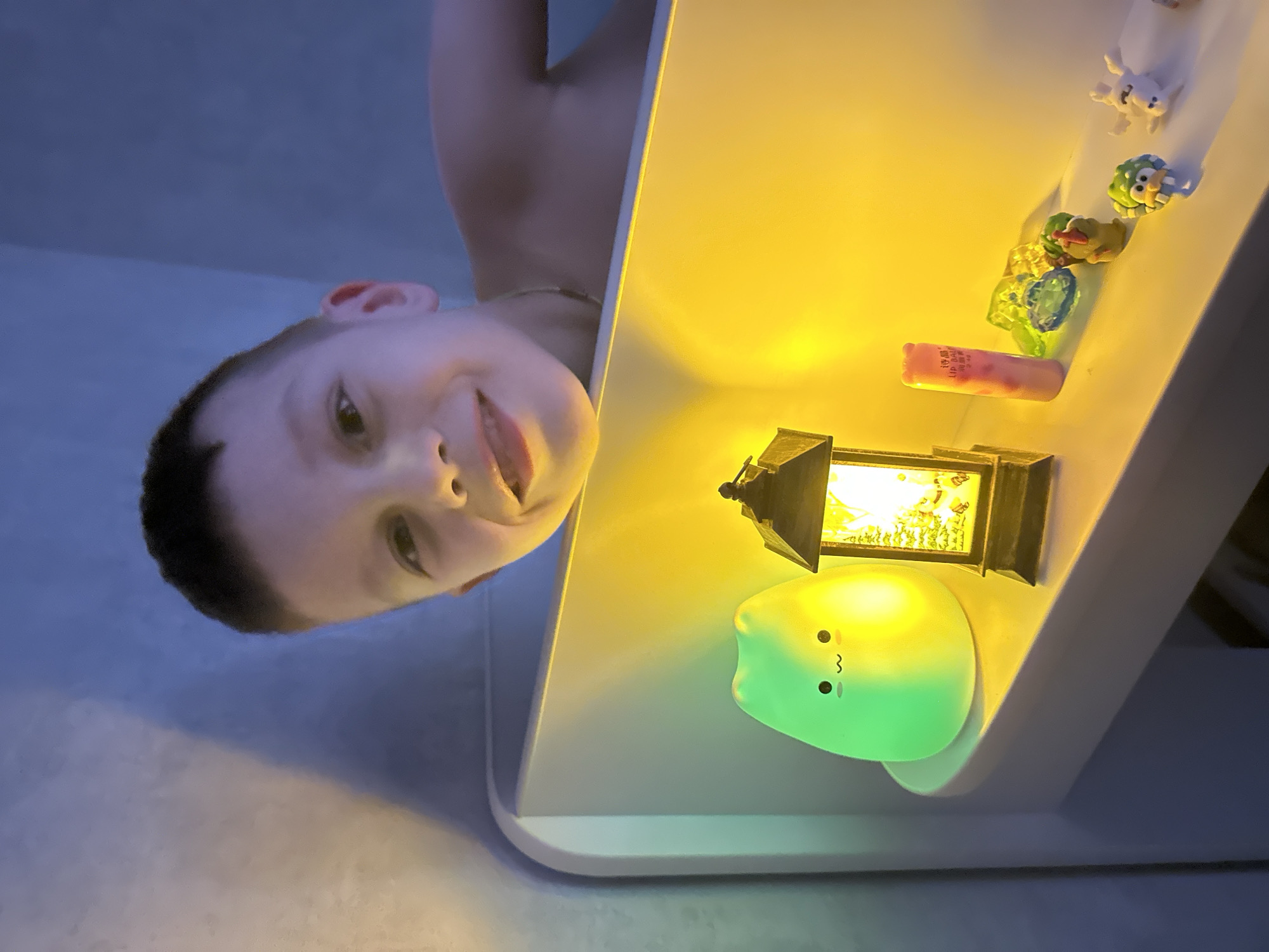 The image, taken in Tomilino, Russia, captures a young boy looking upwards with a wide, joyful smile towards a brightly lit table or shelf. The original photograph is presented from an inverted, low-angle perspective, showing the boy seemingly lying beneath the surface and gazing up at the objects.
The boy, featuring short dark hair and fair skin, displays a happy expression with his mouth open, revealing his teeth. His gaze is directed towards the illuminated items arranged on the light-colored surface above him.
On this table, a collection of objects is bathed in artificial light. A small, dark-toned decorative lantern casts a warm yellow glow, illuminating a miniature scene within its glass panes. Adjacent to it is a soft, glowing, light-green blob-shaped lamp, characterized by small black eyes and a wavy mouth, resembling a playful creature. Further along, a pink cylindrical item, identifiable as lip balm, has partially visible text including Chinese characters '唇晶' (chún jīng, meaning 'lip crystal' or 'lip balm') and the English word 'LIP'. Alongside these are several small, colorful toy figures: a white bunny-like figure, a green spiky creature, a yellow duck-like figure with a red hat, and multiple clear, faceted jewel-like objects in shades of green, blue, and orange.
The background surrounding the boy is a soft, muted blue-gray, suggesting dim ambient lighting in the room, indicative of evening or nighttime. The strong, localized illumination from the lamps on the table creates a striking contrast, casting vibrant yellow and green light onto the objects. The scene evokes a sense of wonder and playful engagement as the child observes the illuminated display.