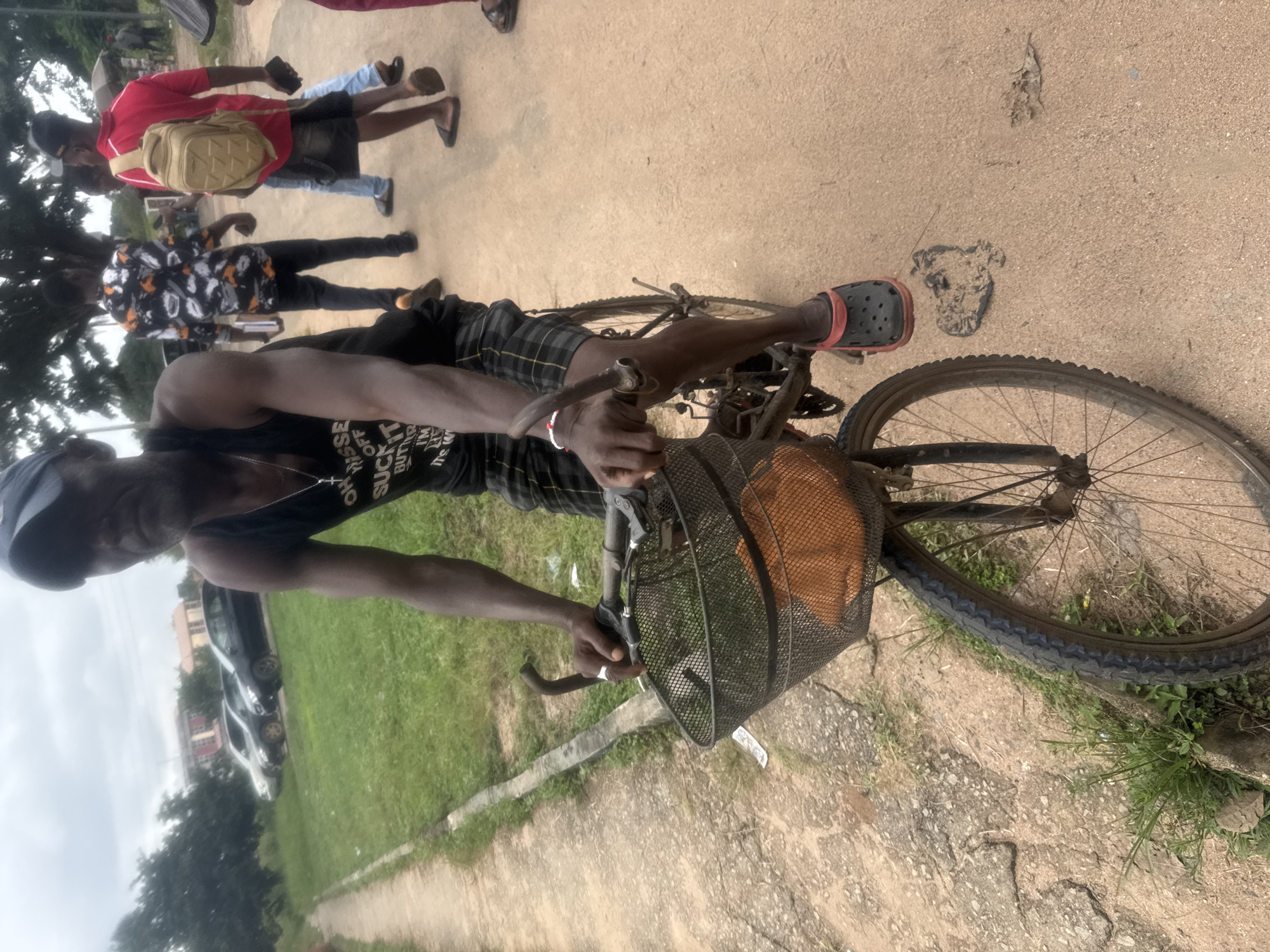 A Black man wearing a dark blue baseball cap, a black tank top with white lettering that reads "OH YES SUCK IT BUTTERFLY I'M SO HIGH IT'S WILD", and checkered shorts rides a bicycle on a dirt and gravel road. The bicycle has a wire basket on the front containing an orange object. The man's right hand is on the handlebars, and his left hand is also on the handlebars. He is wearing a red and black sandal on his right foot. In the background, several people are walking. One man wears a red shirt and a beige backpack. Another man wears a black and white patterned shirt. Two cars are parked on the left side of the road, near a grassy area and some trees. The sky is overcast. The scene appears to be in Ihiagwa, Nigeria.